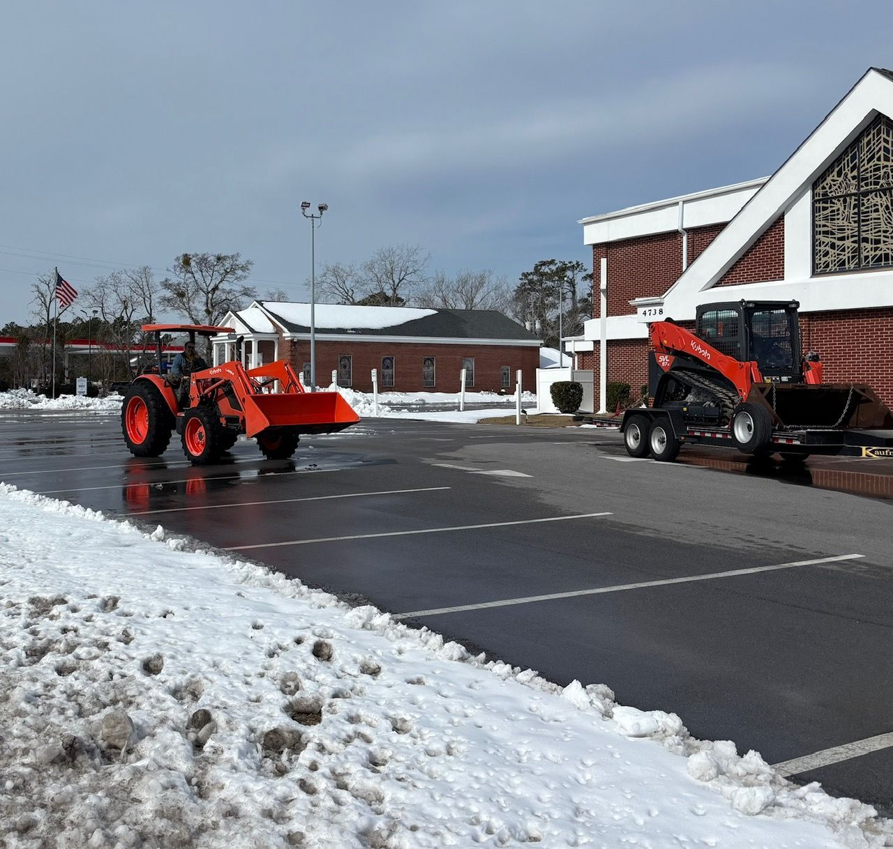 An orange tractor and a skid steer parked on a snow-covered parking lot in front of a brick building.
