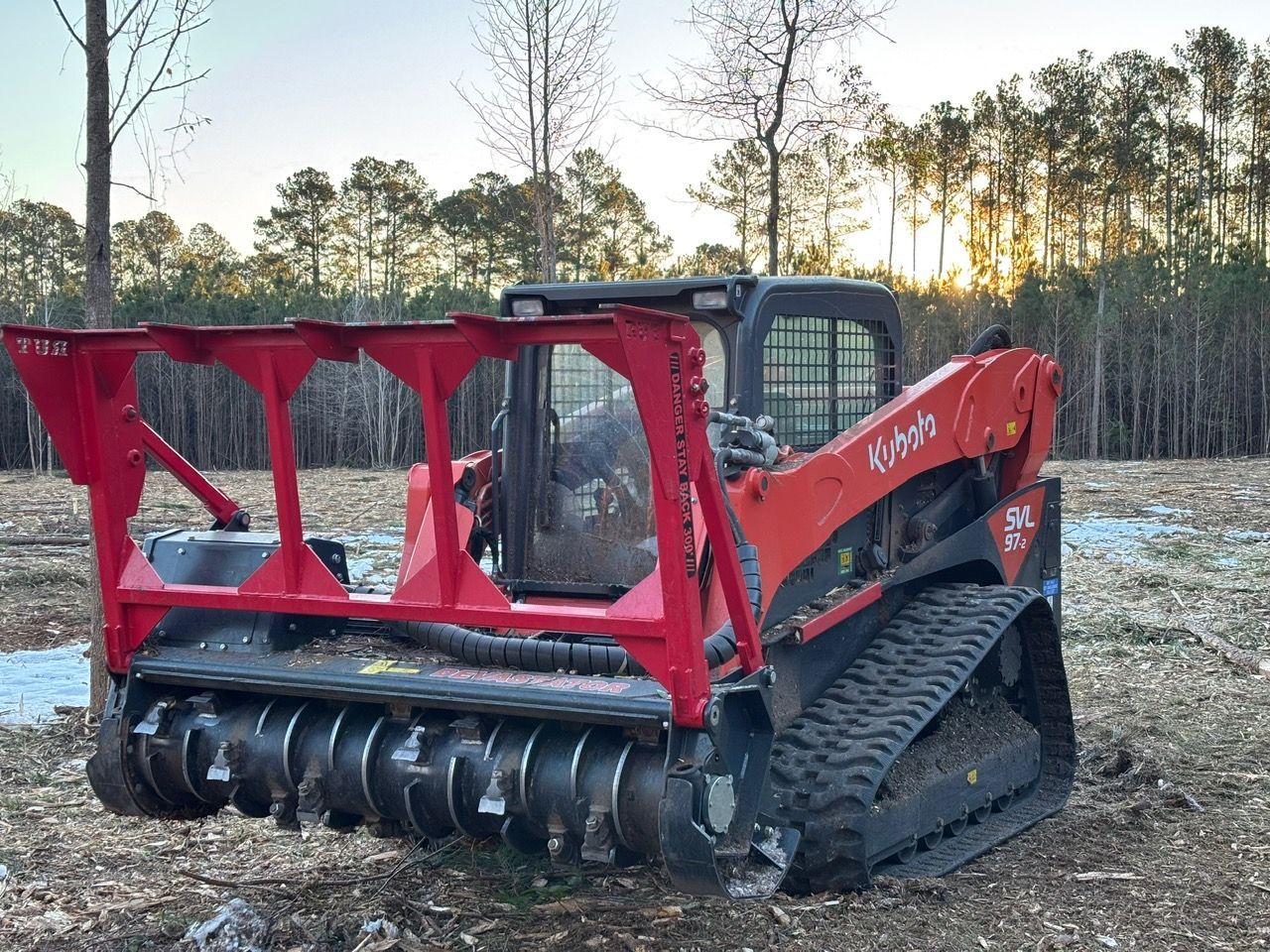 A red Kubota compact track loader equipped with a forestry mulcher attachment sits in a cleared wooded area at sunset.