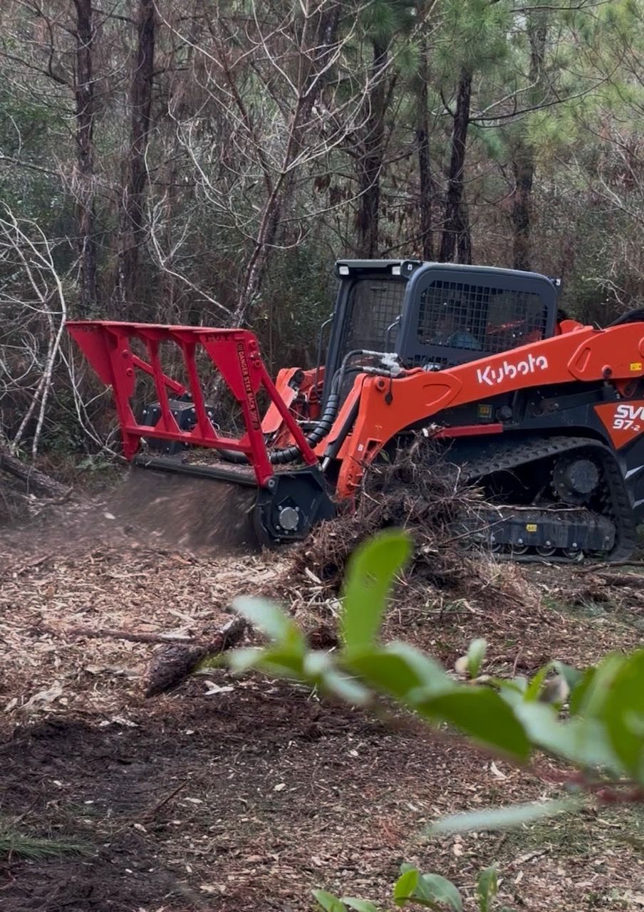 An orange Kubota track loader with a red root rake attachment clearing brush in a wooded area.