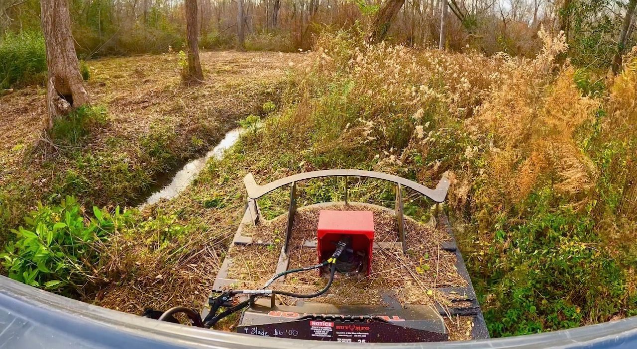 View from inside a cab looking at a mower cutting grass and brush near a small drainage ditch in a wooded area.