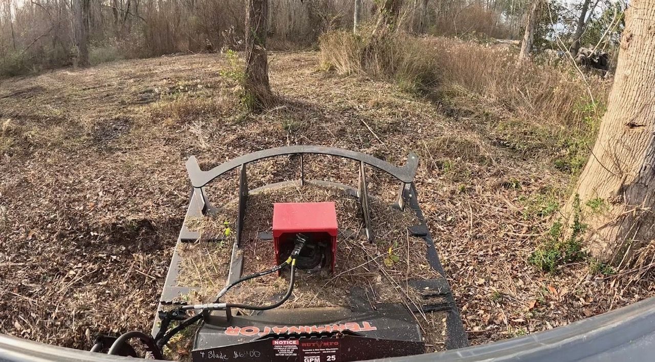 View from a tractor cab looking over a brush cutter attachment resting on leaf-covered ground in a wooded area.