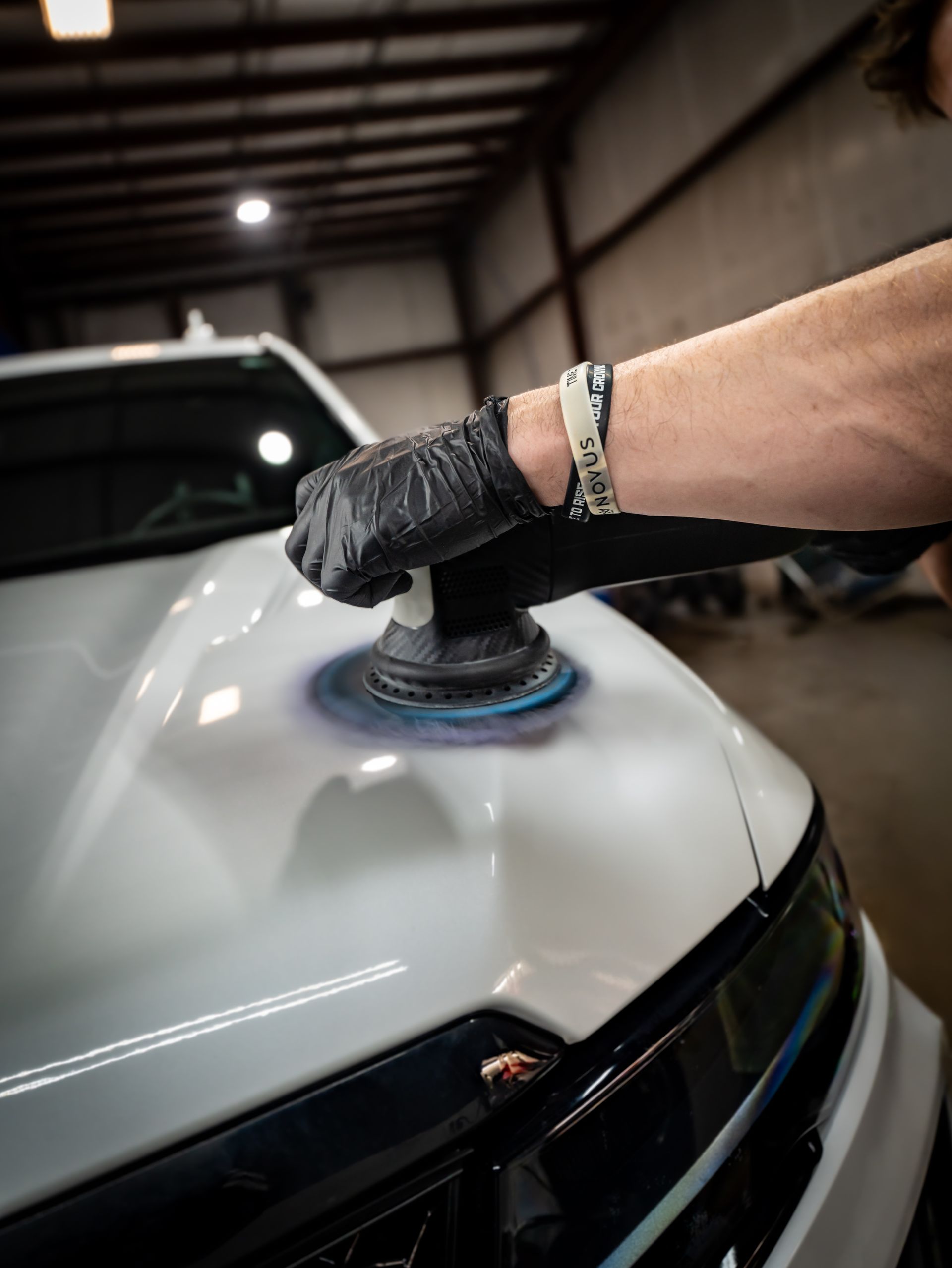 Gloved hand polishing a white car hood with a buffer in a garage