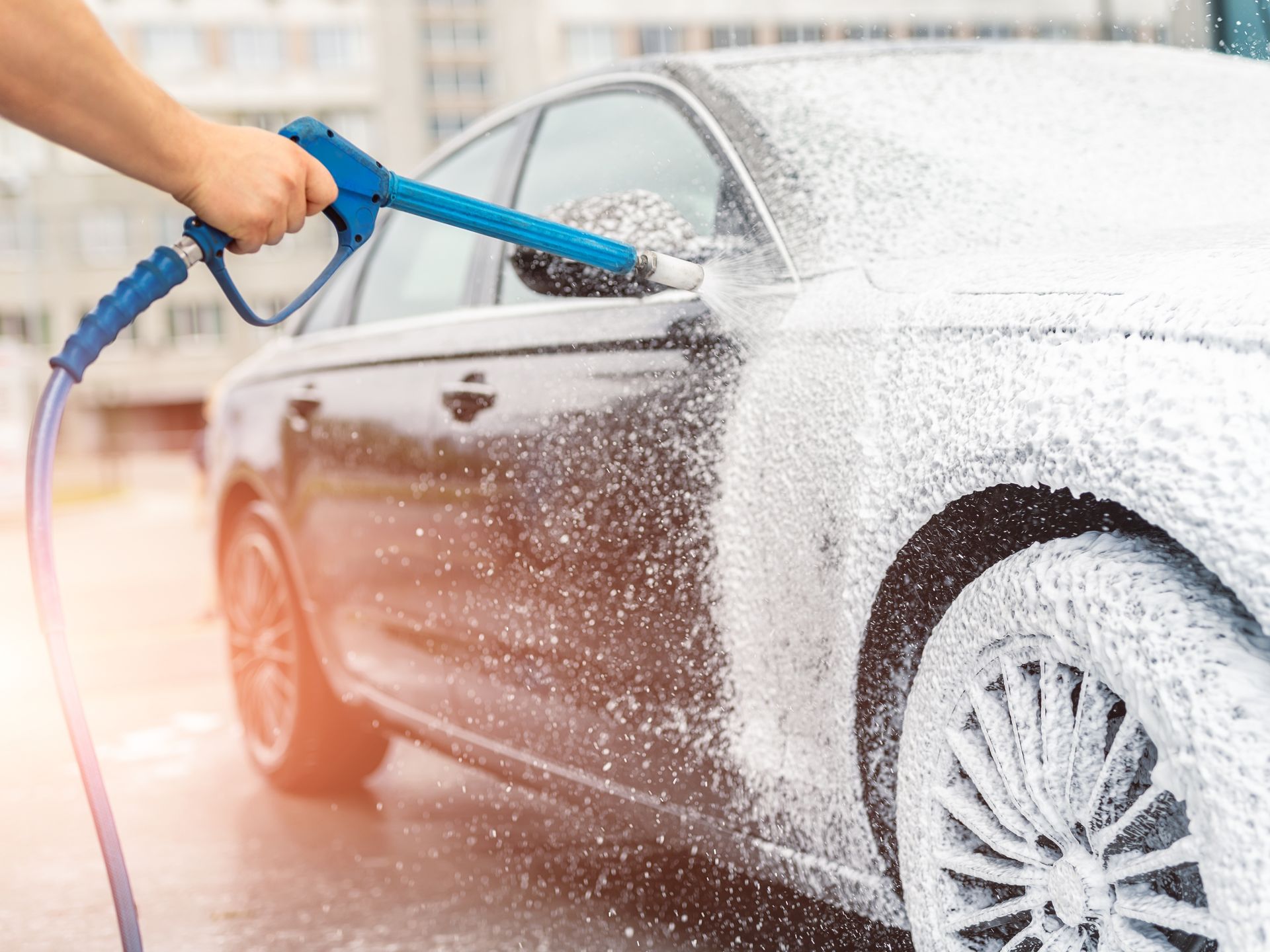 Hand washing a silver car with a blue hose, spraying soap and water onto the front wheel and door
