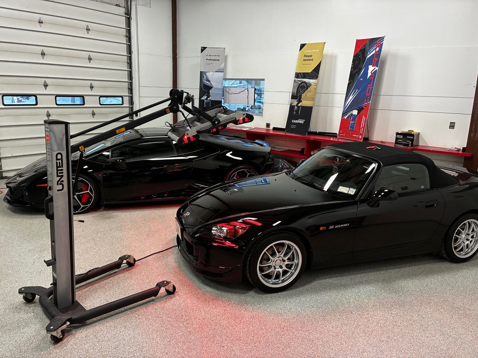 Two black sports cars in a garage, one on a lift, with automotive equipment and panels nearby