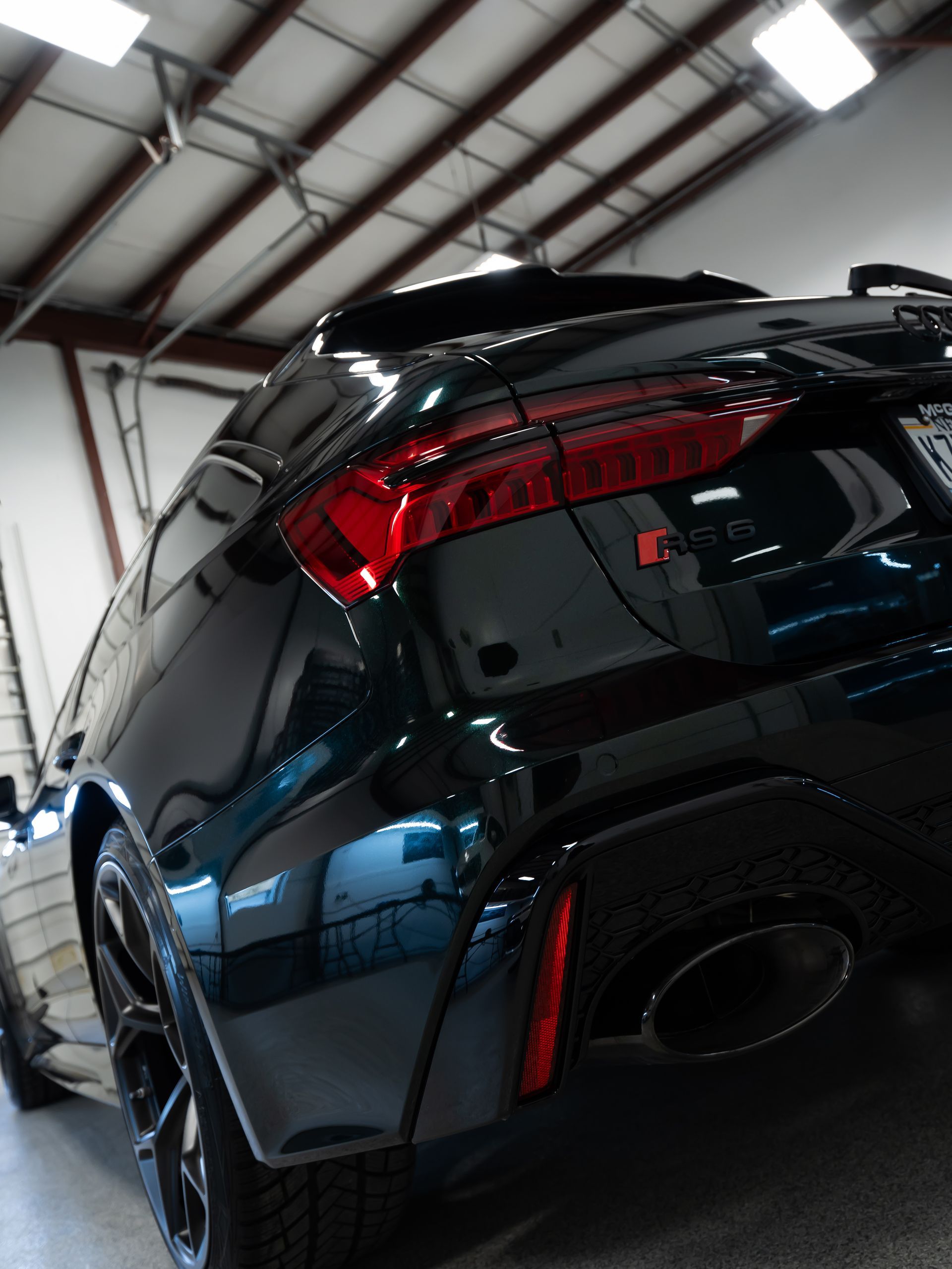 Glossy black sports car rear in a garage, with red taillight and ceiling lights reflecting on the body