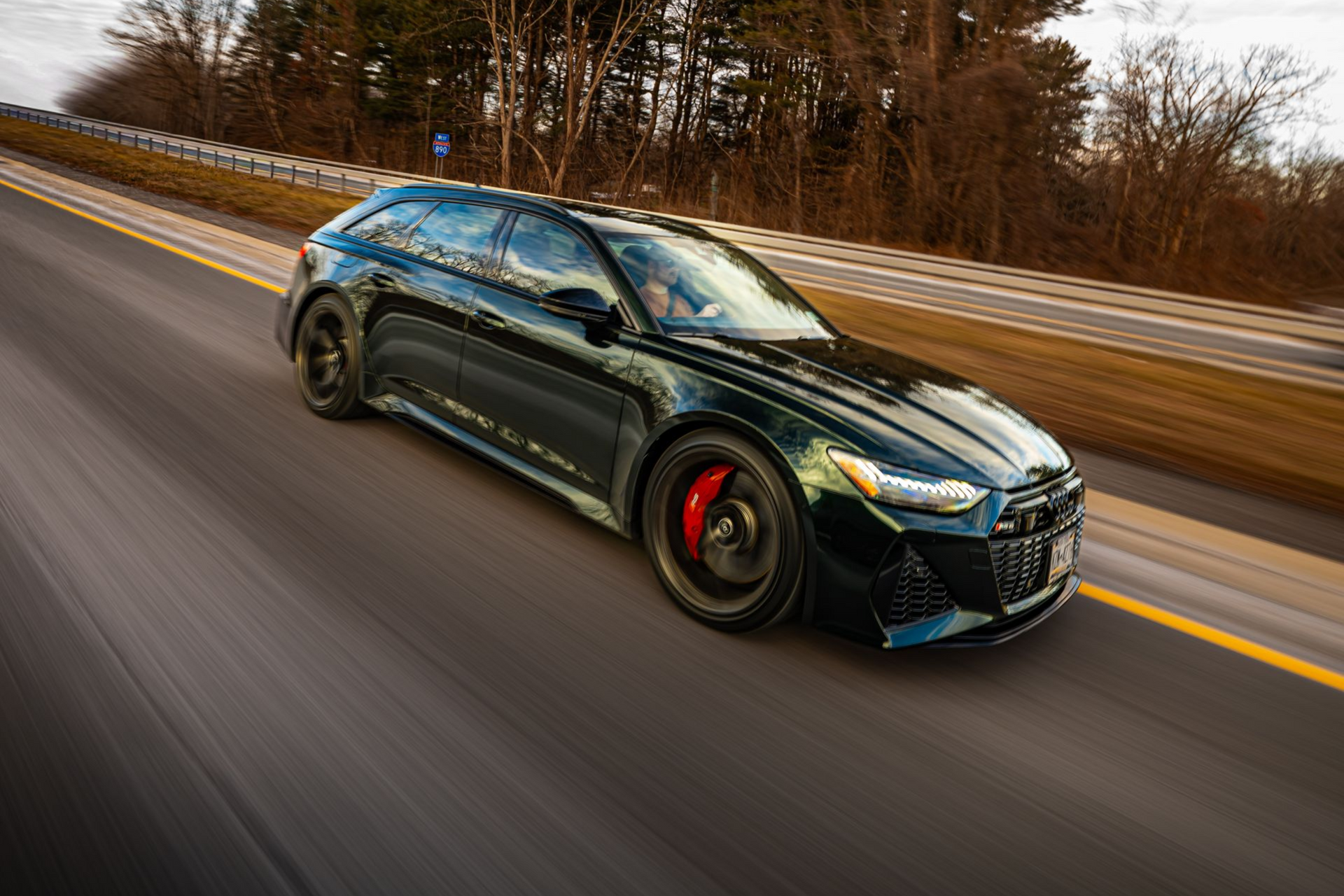 Black sports hatchback speeding on a wet road, blurred trees and guardrail in the background