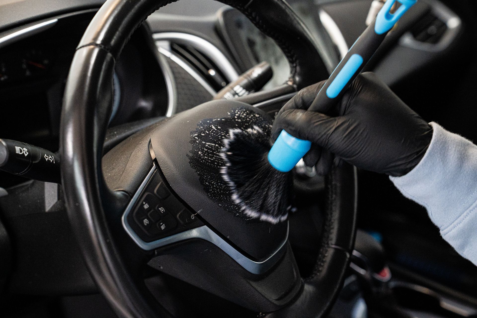 Gloved hand cleaning a car steering wheel with a brush and foam.