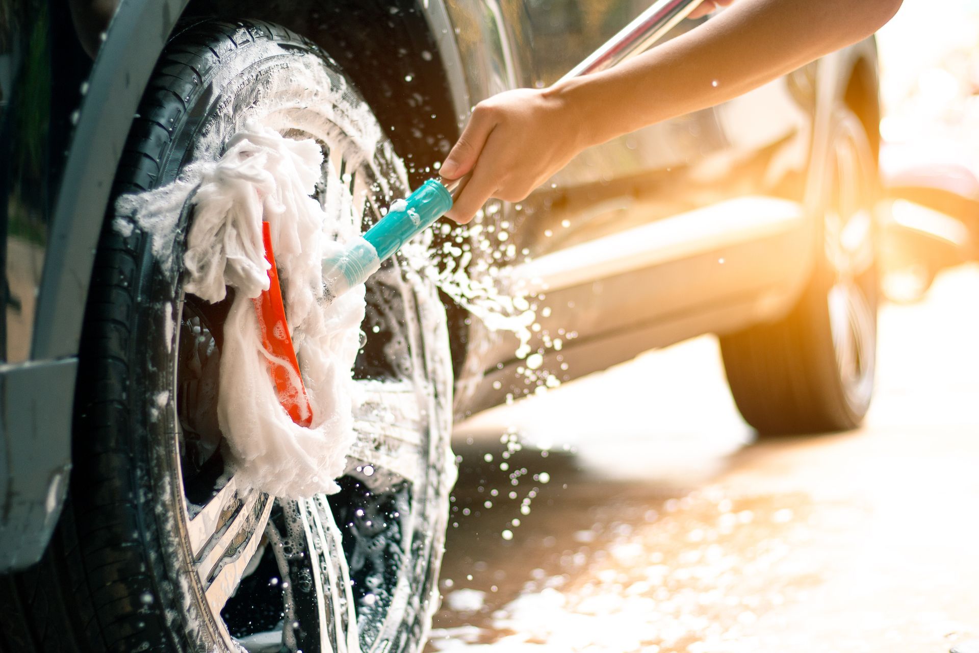 Person washing a car wheel with soap and water in bright sunlight