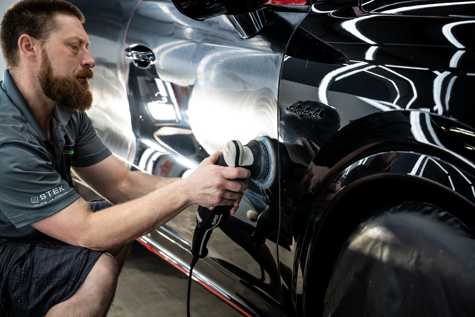 Detailer polishing a black car with a buffer in a garage