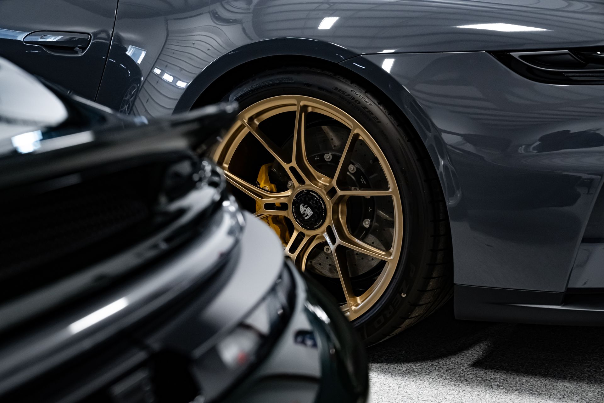 Close-up of two luxury sports cars, with gold rims on a dark gray car in a garage.
