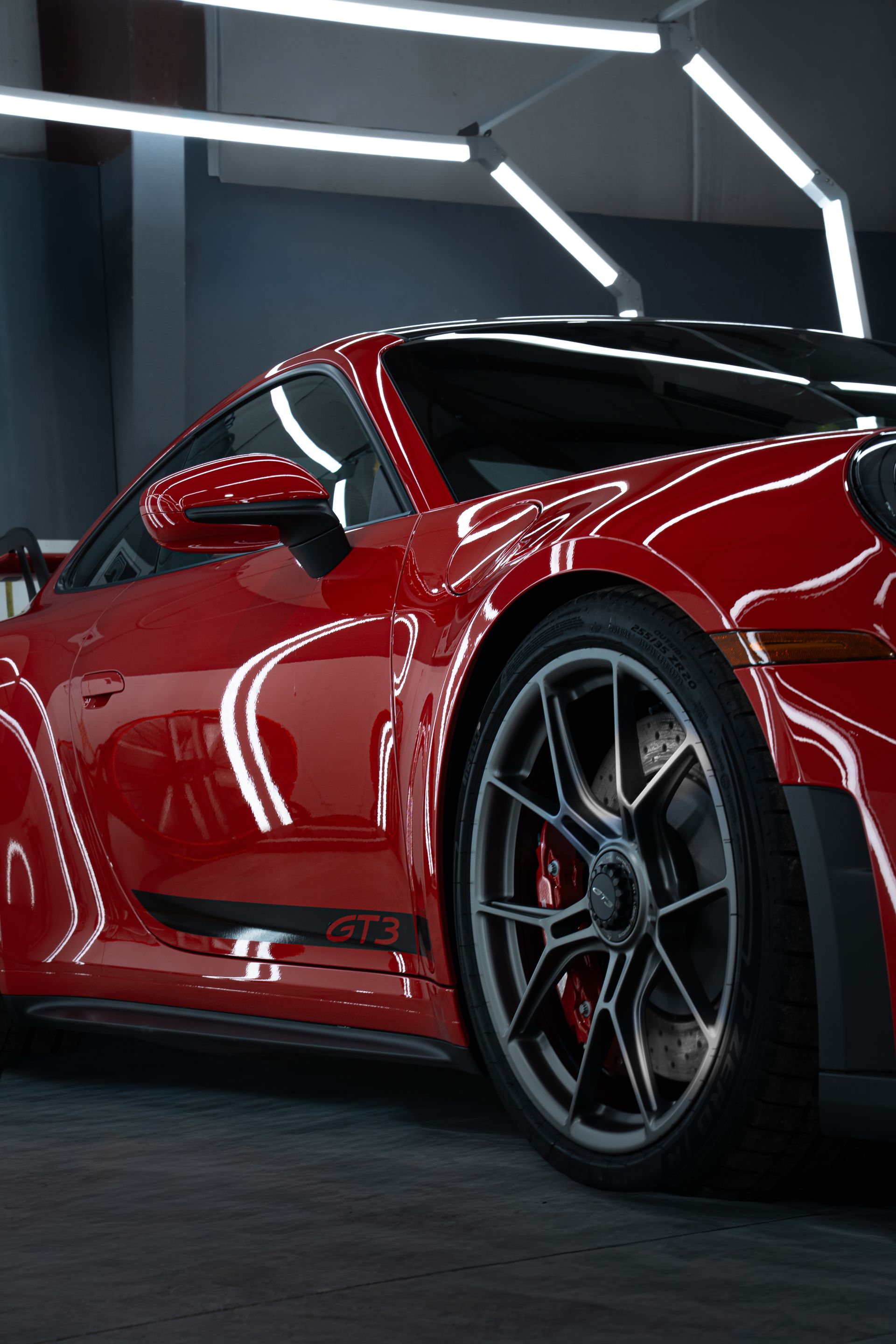 Red sports car in a dark showroom, shown from the side with bright overhead lights.