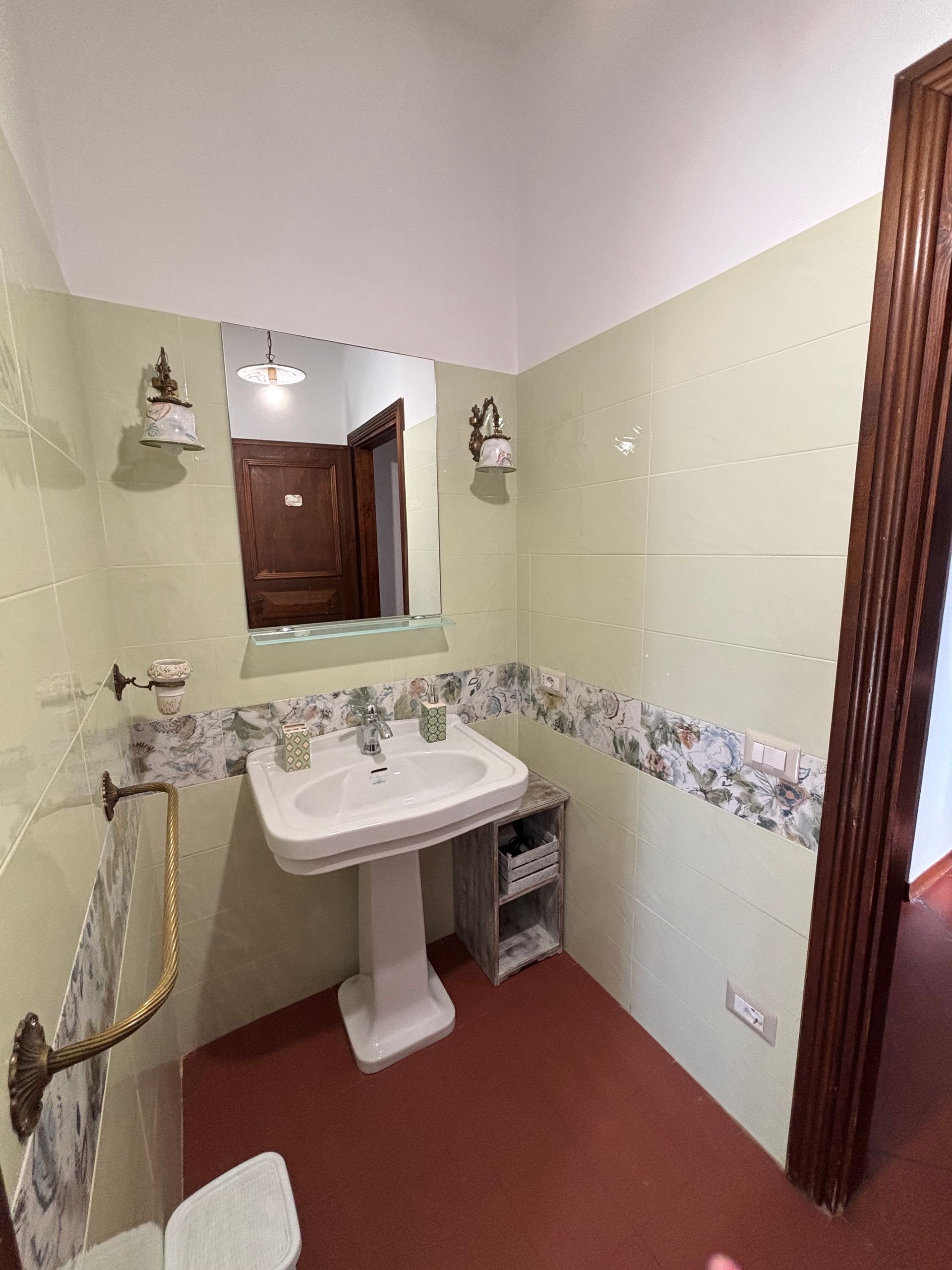 Bagno con lavabo a colonna, specchio, applique e pavimento in piastrelle rosse. Pareti verdi, stipite della porta in legno.