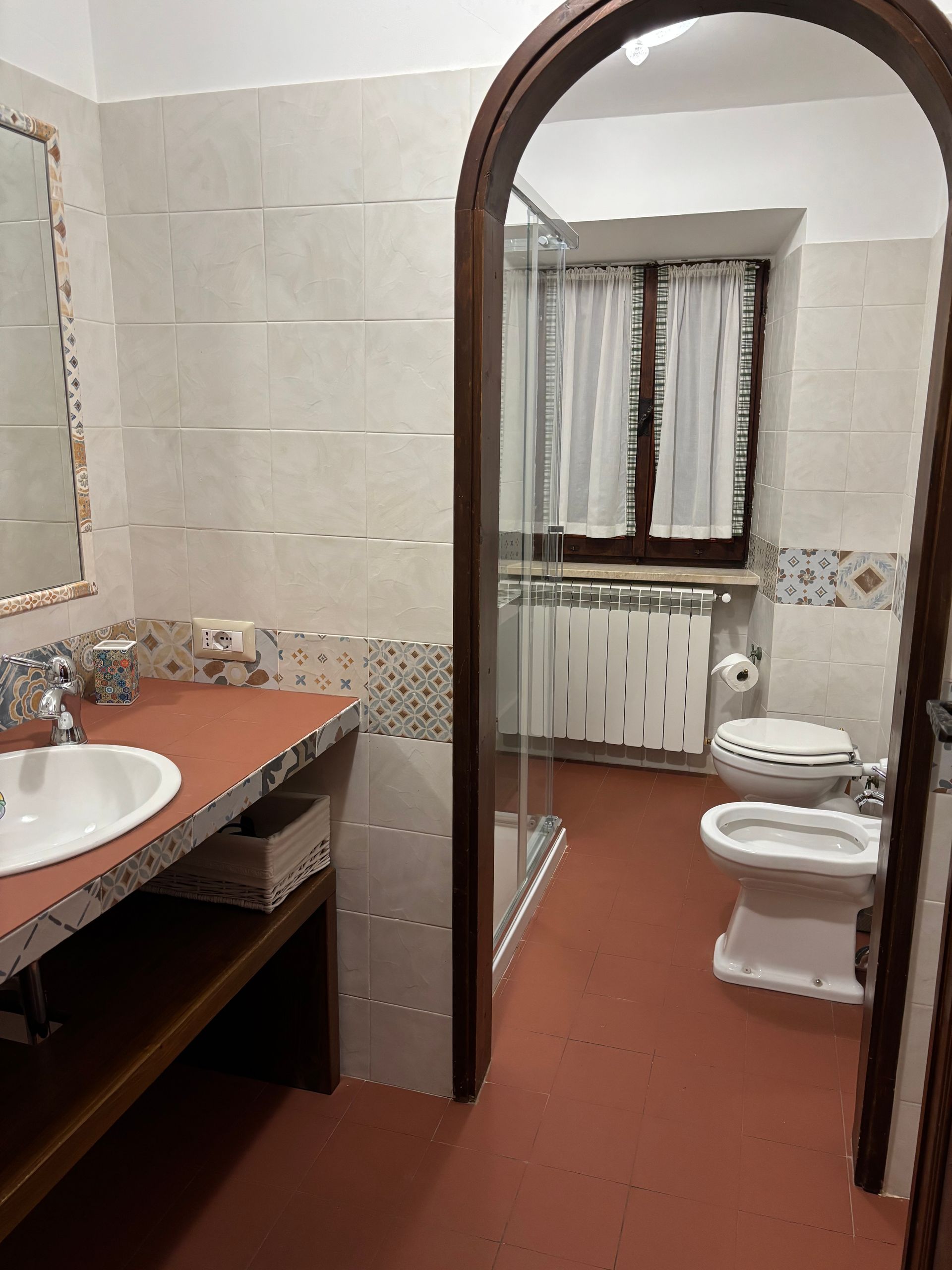 Bagno con pavimento rosso, lavandino, WC, bidet, doccia e finestra con tende.