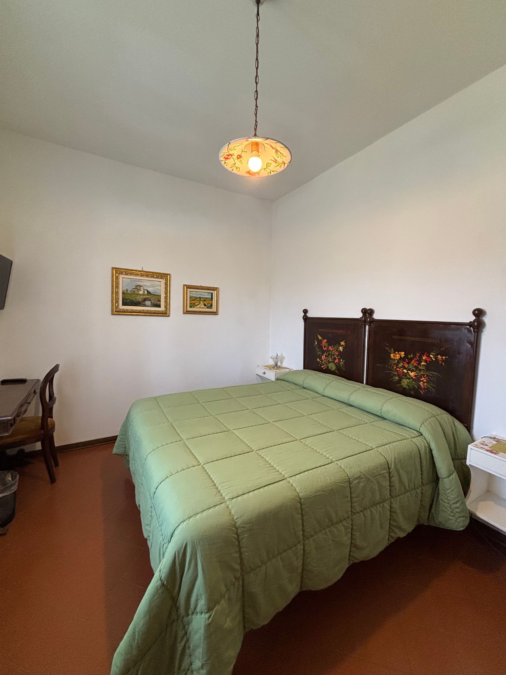 Camera da letto con letto, quadri, scrivania e lampada a sospensione. Pareti bianche, trapunta verde e pavimento in terracotta.