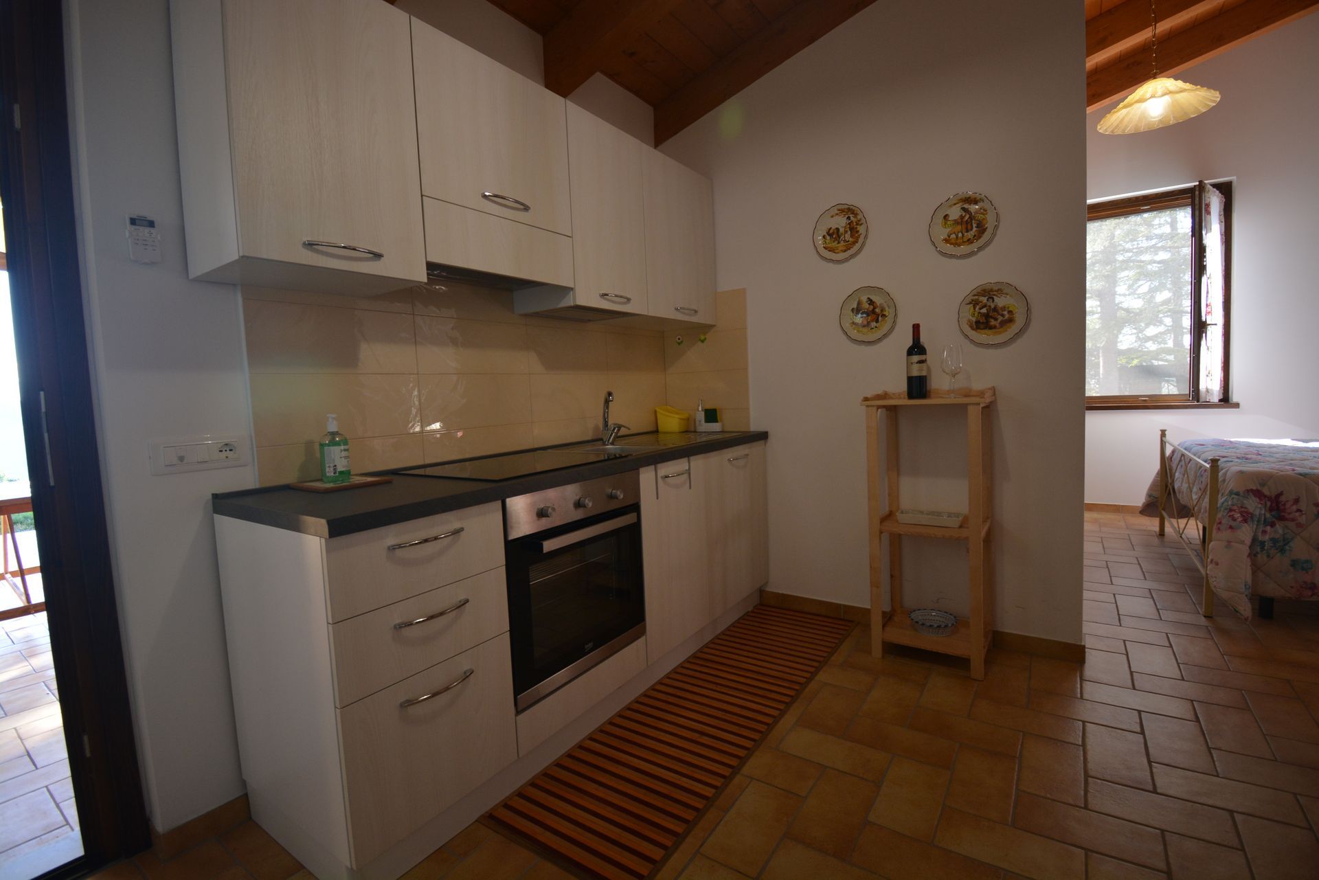 Cucina con mobili bianchi, forno e piccolo tappeto, adiacente alla camera da letto con letto e finestra.