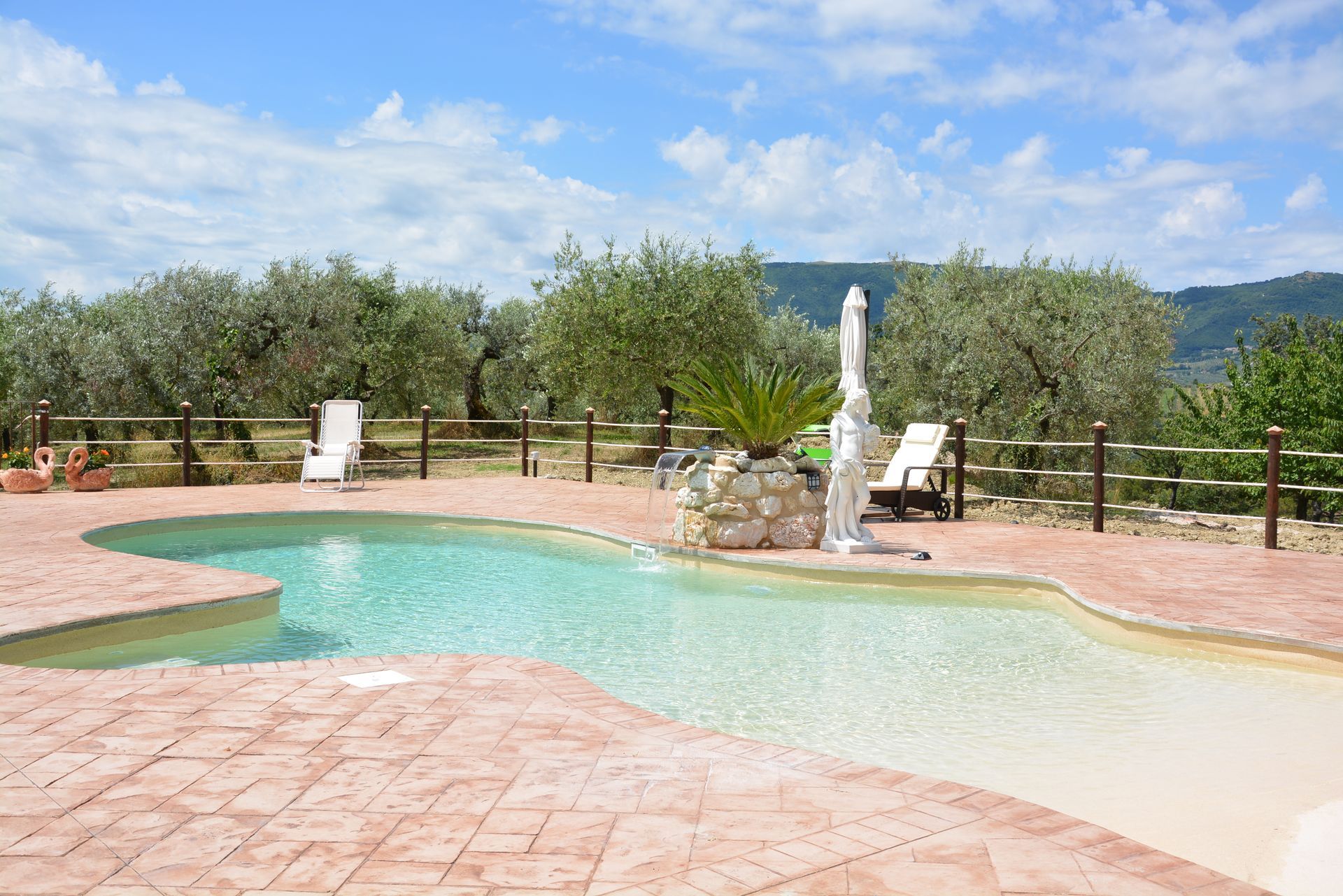 Piscina con fontana, circondata da alberi e un patio in mattoni sotto un cielo azzurro.