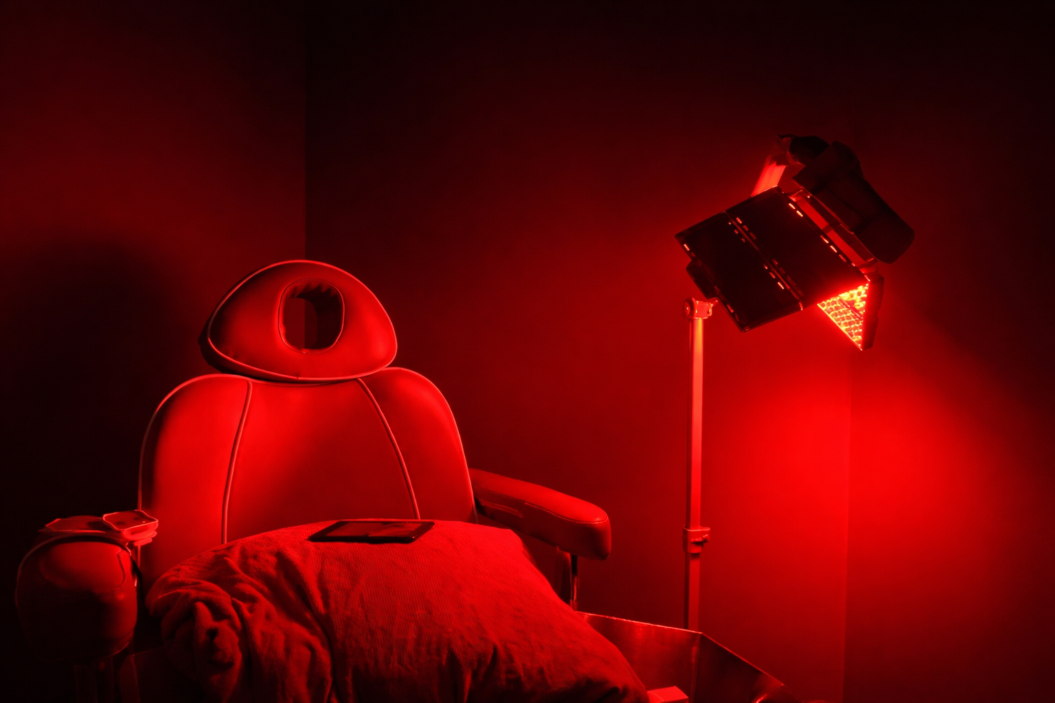 A medical treatment chair bathed in bright red light, positioned next to a tall, professional lamp in a dark room.