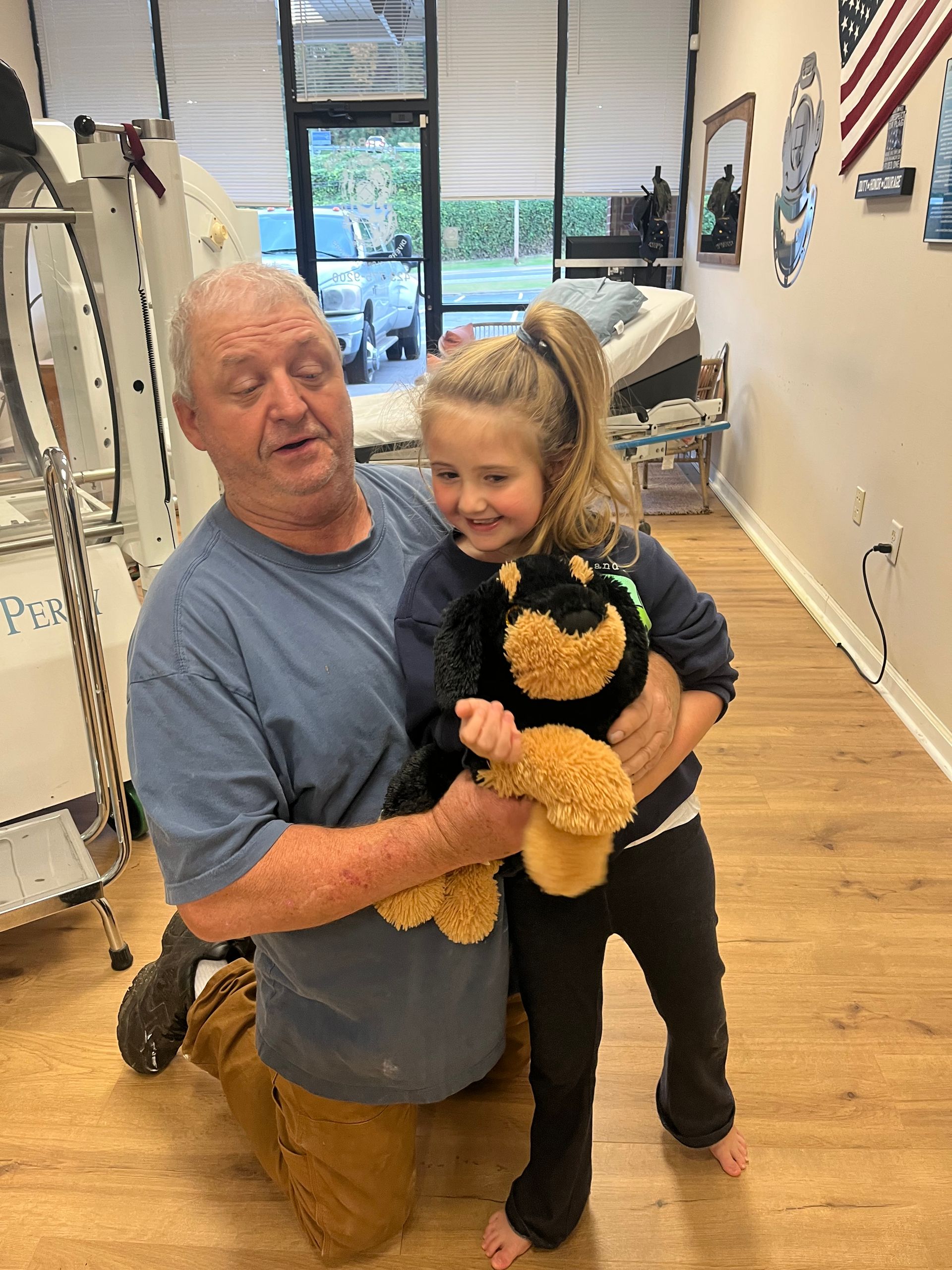 Man kneels beside smiling child holding a teddy bear in a bright room.