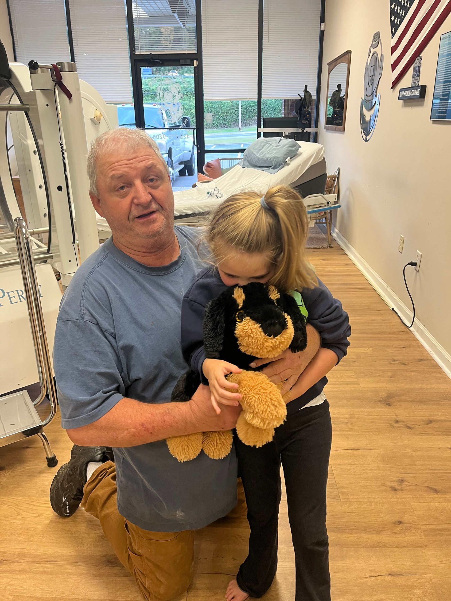 An older man kneels beside a child holding a stuffed bear in a bright room with wooden floors.