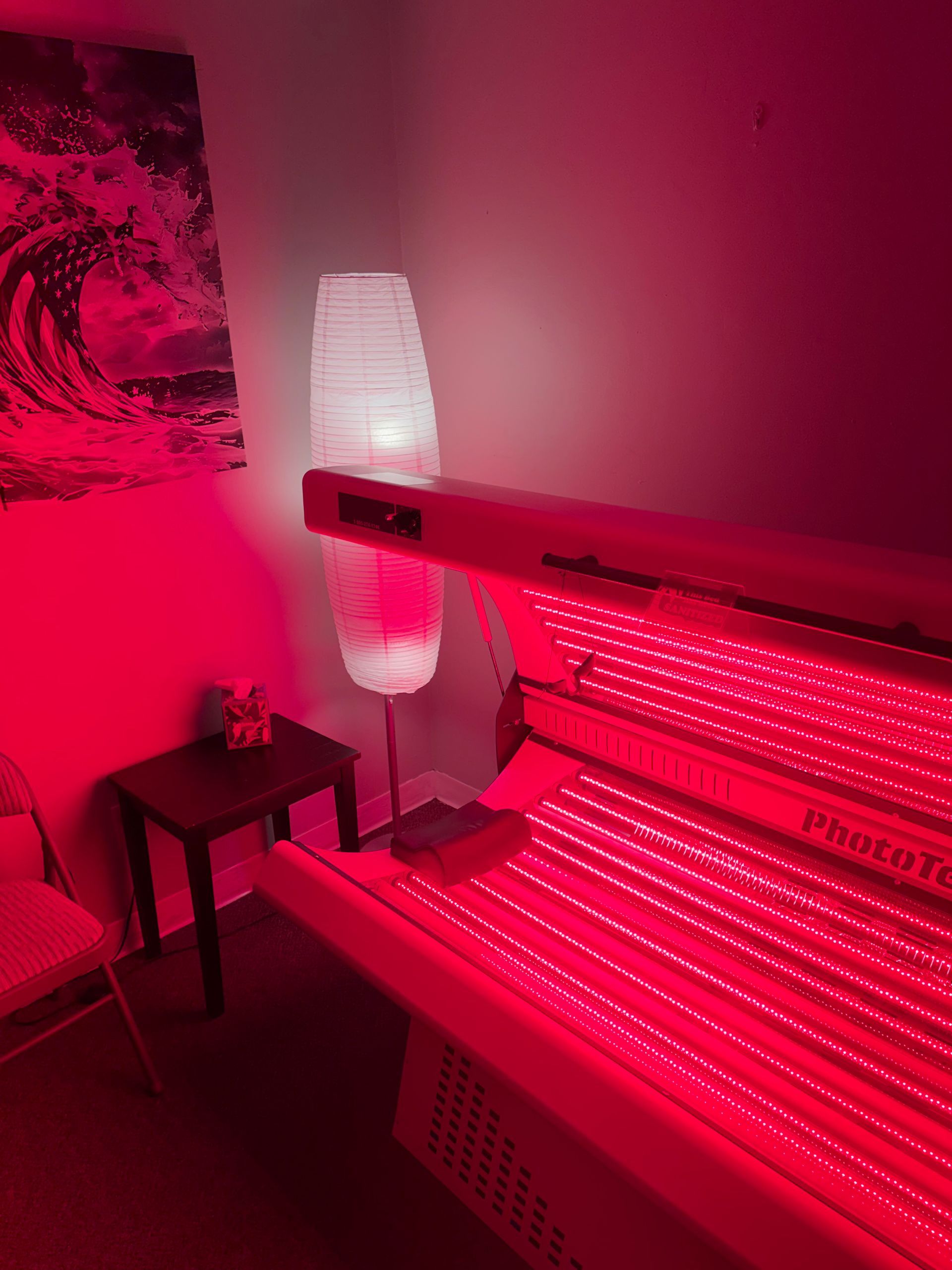 Red-lit room with a tanning bed, standing lamp, and small side table by a wall poster.