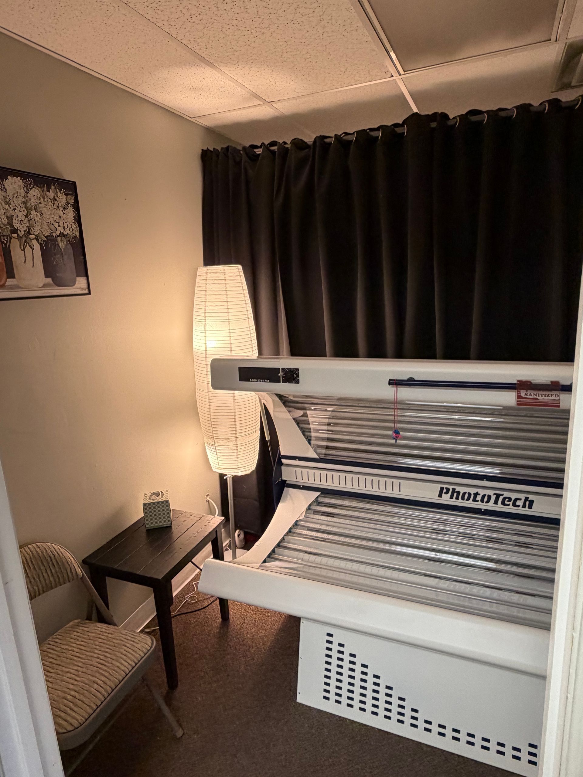 Cozy room with a tanning bed, floor lamp, small table, chair, and dark curtains.
