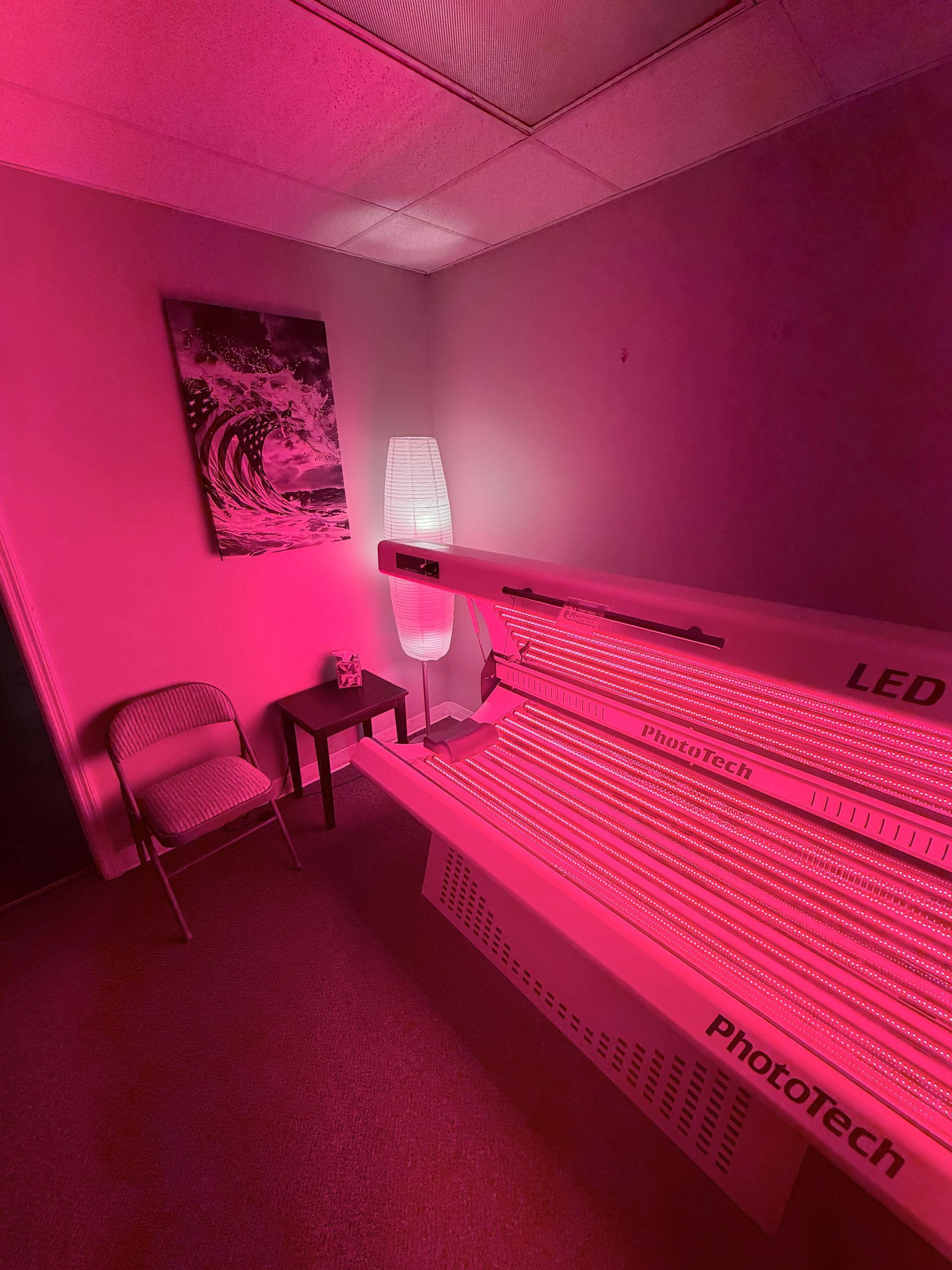A small room bathed in red light, featuring a Phatotech LED light therapy bed, a folding chair, and wall artwork.