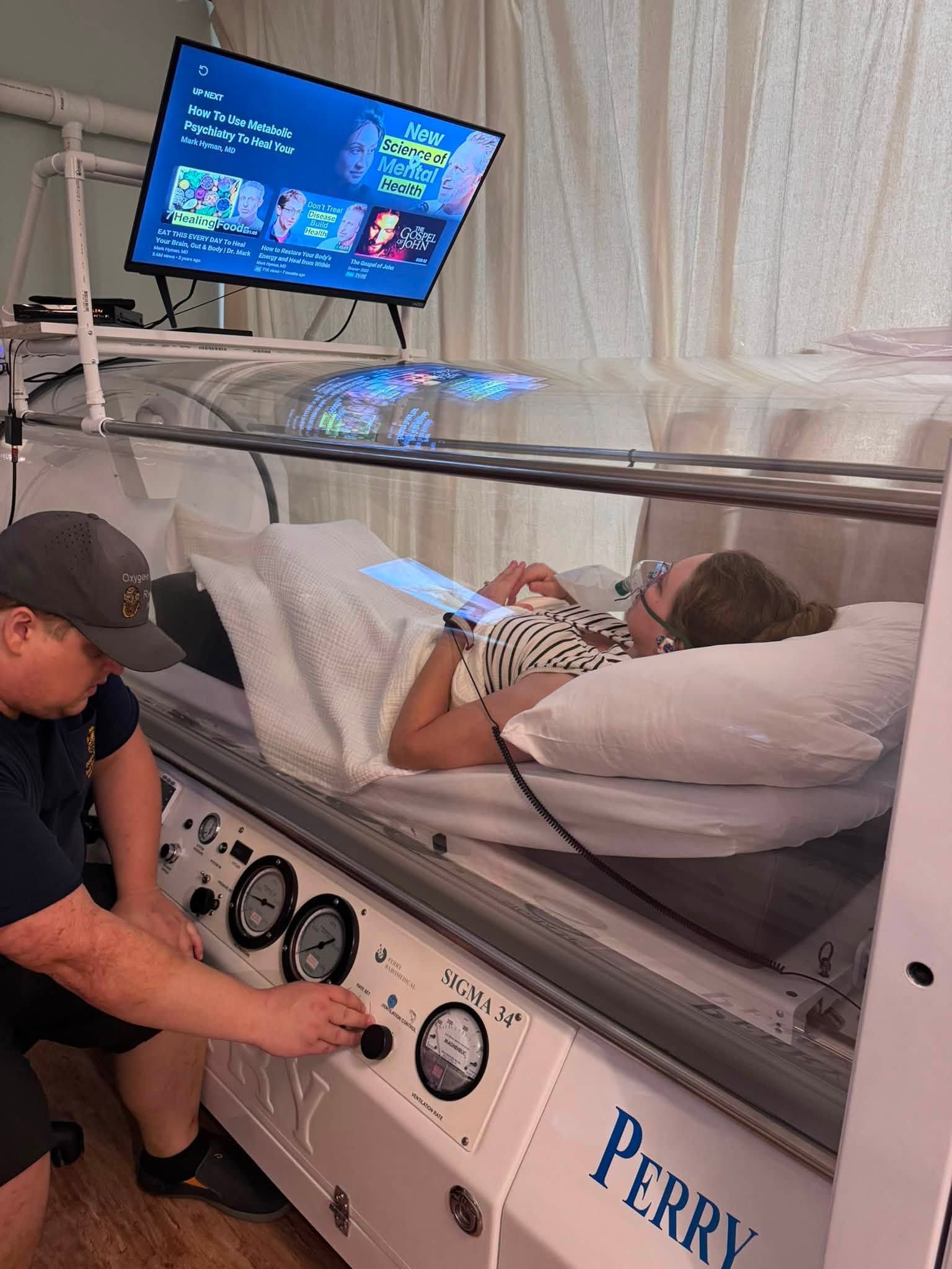 A technician monitors a patient inside a hyperbaric oxygen therapy chamber in a clinical setting with blue lighting.