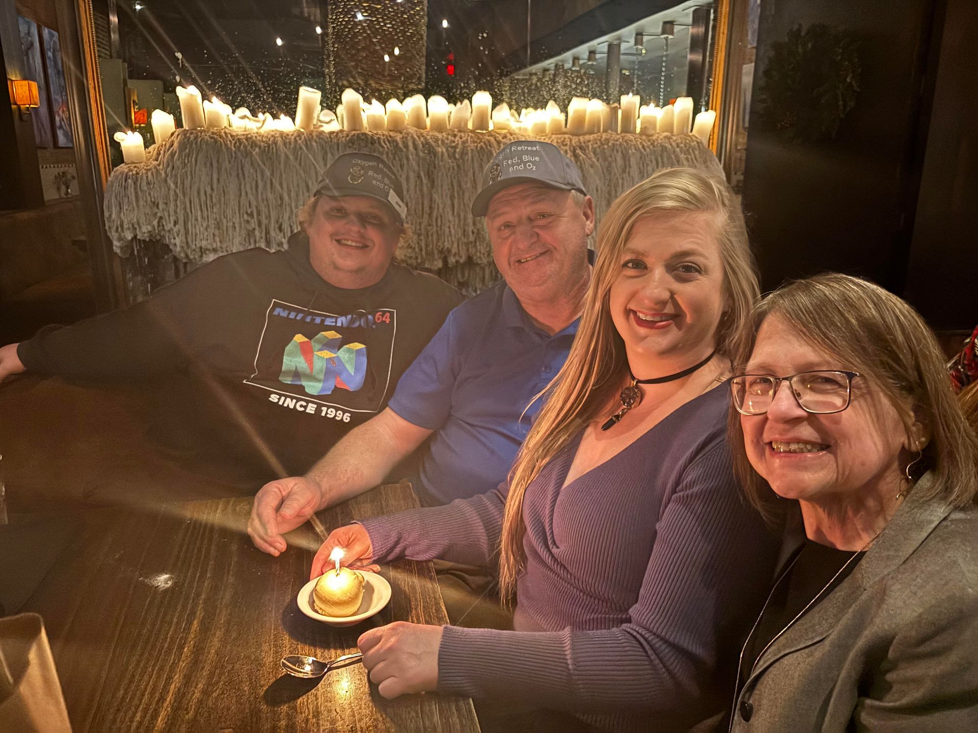 Four people seated at a restaurant table, smiling with a lit dessert in the foreground.