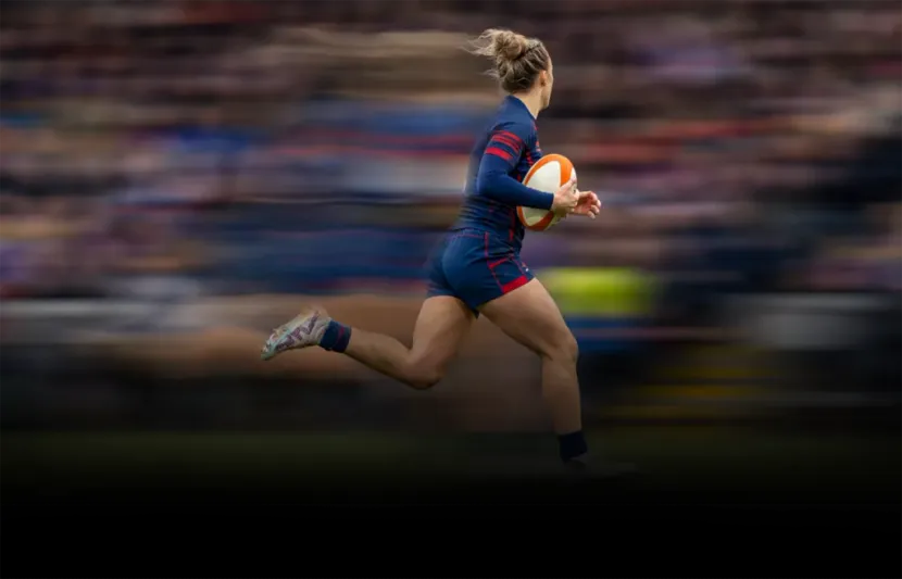 Rugby player in navy and red sprints with the ball across the field, motion blurred background
