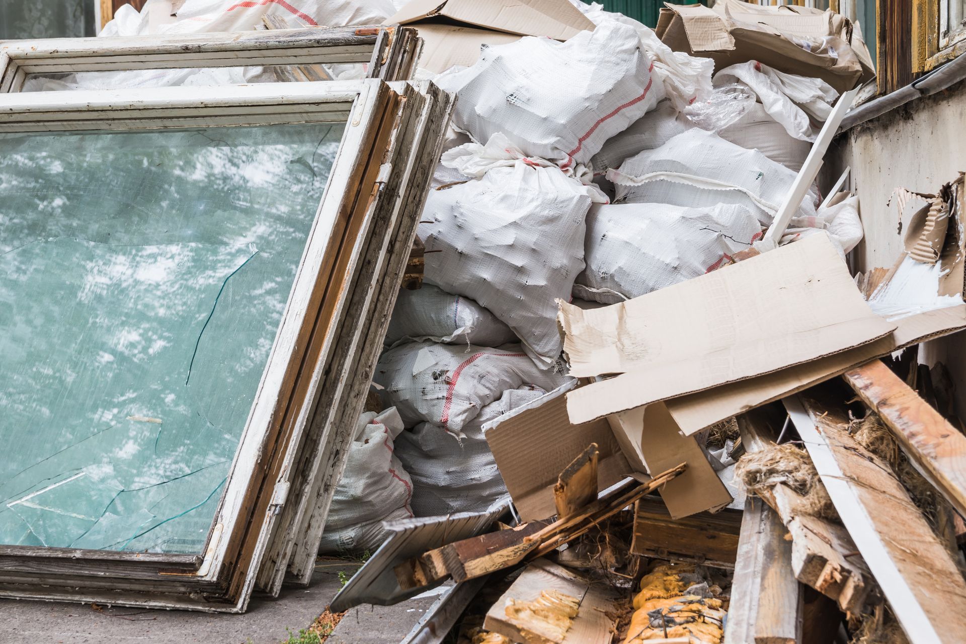 A pile of construction debris featuring old window frames, cardboard, and white waste sacks.