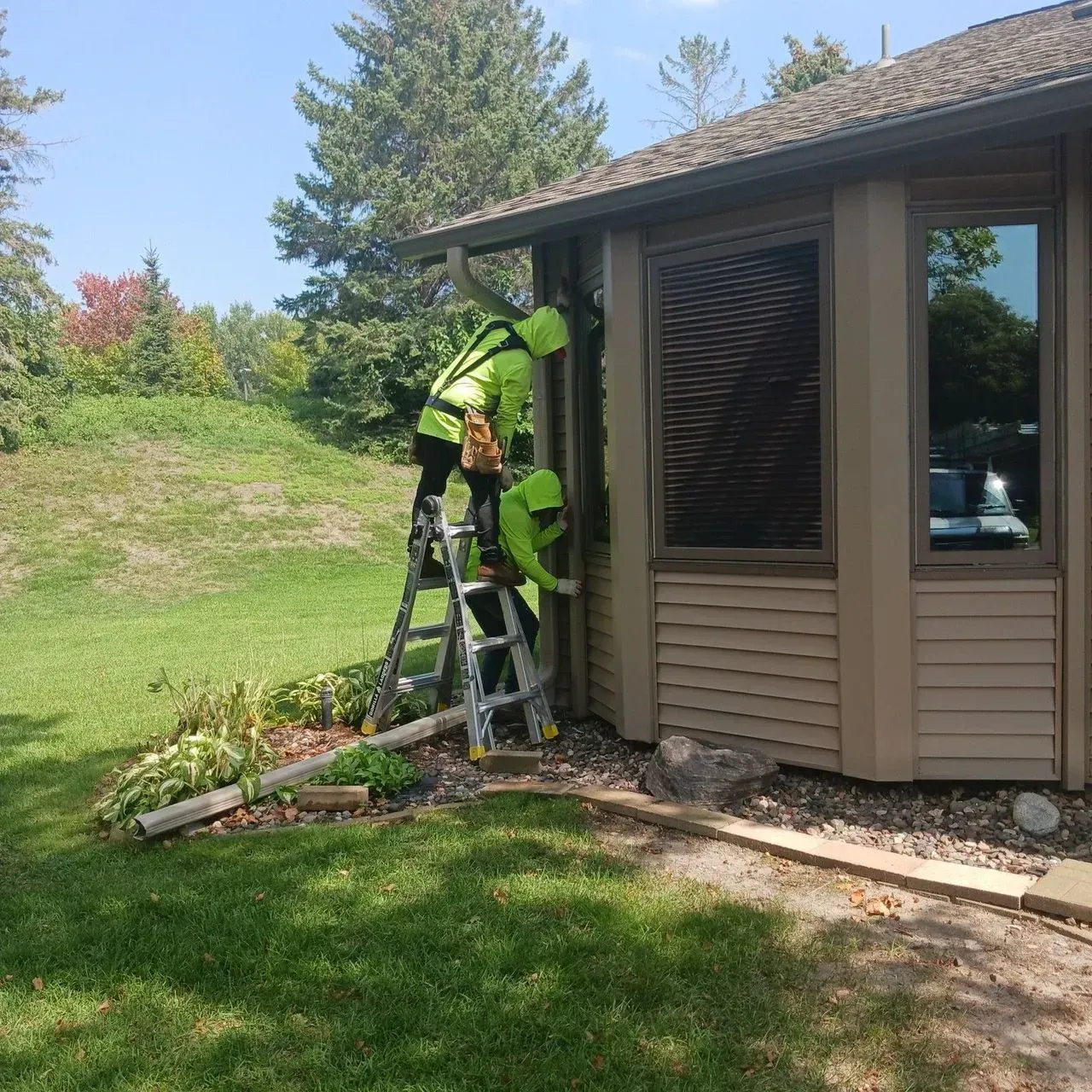 Two workers in neon vests on a ladder cleaning a gutter on a house with brown siding.