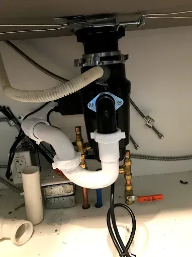 Under-sink plumbing with white drain pipes, black disposer, and exposed hoses on a kitchen cabinet floor