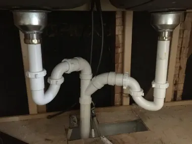 Under-sink plumbing with white PVC pipes beneath two stainless steel sinks