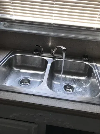 Double kitchen sink with faucet beneath window blinds, stainless steel and black countertop.