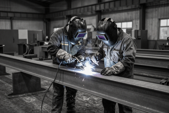 Two welders in protective gear weld a large steel beam in an industrial workshop.