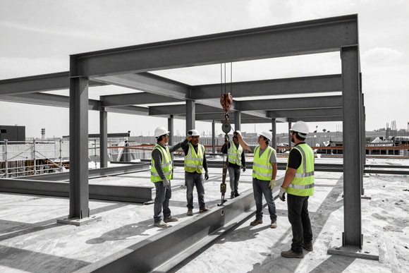 Construction workers in high-visibility vests stand beneath a steel building frame on a site.