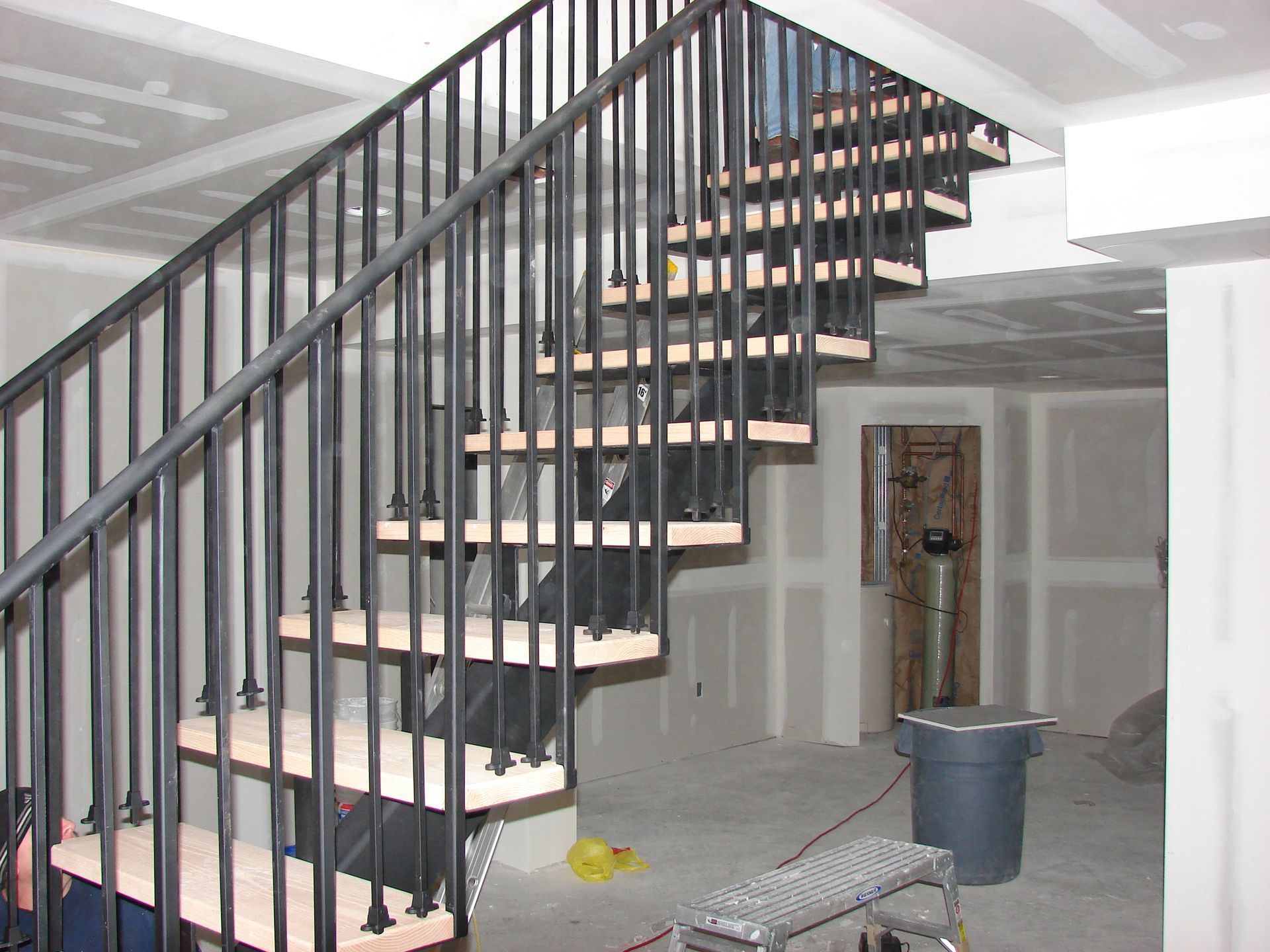 Unfinished interior with a staircase, exposed drywall, a yellow bucket on the floor, and a trash can nearby