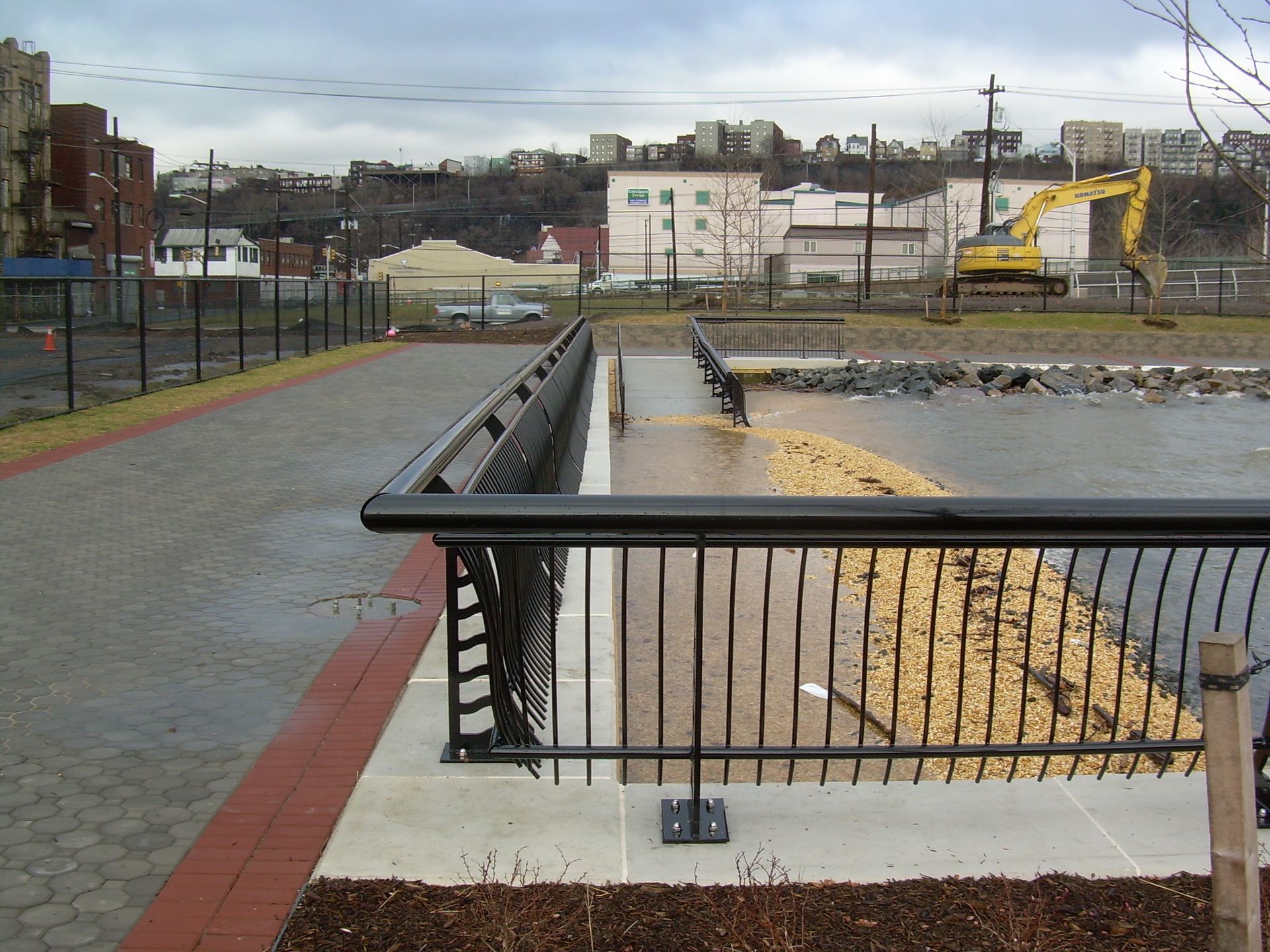 Riverside walkway with black railing beside muddy water, cloudy sky, and construction equipment in the distance