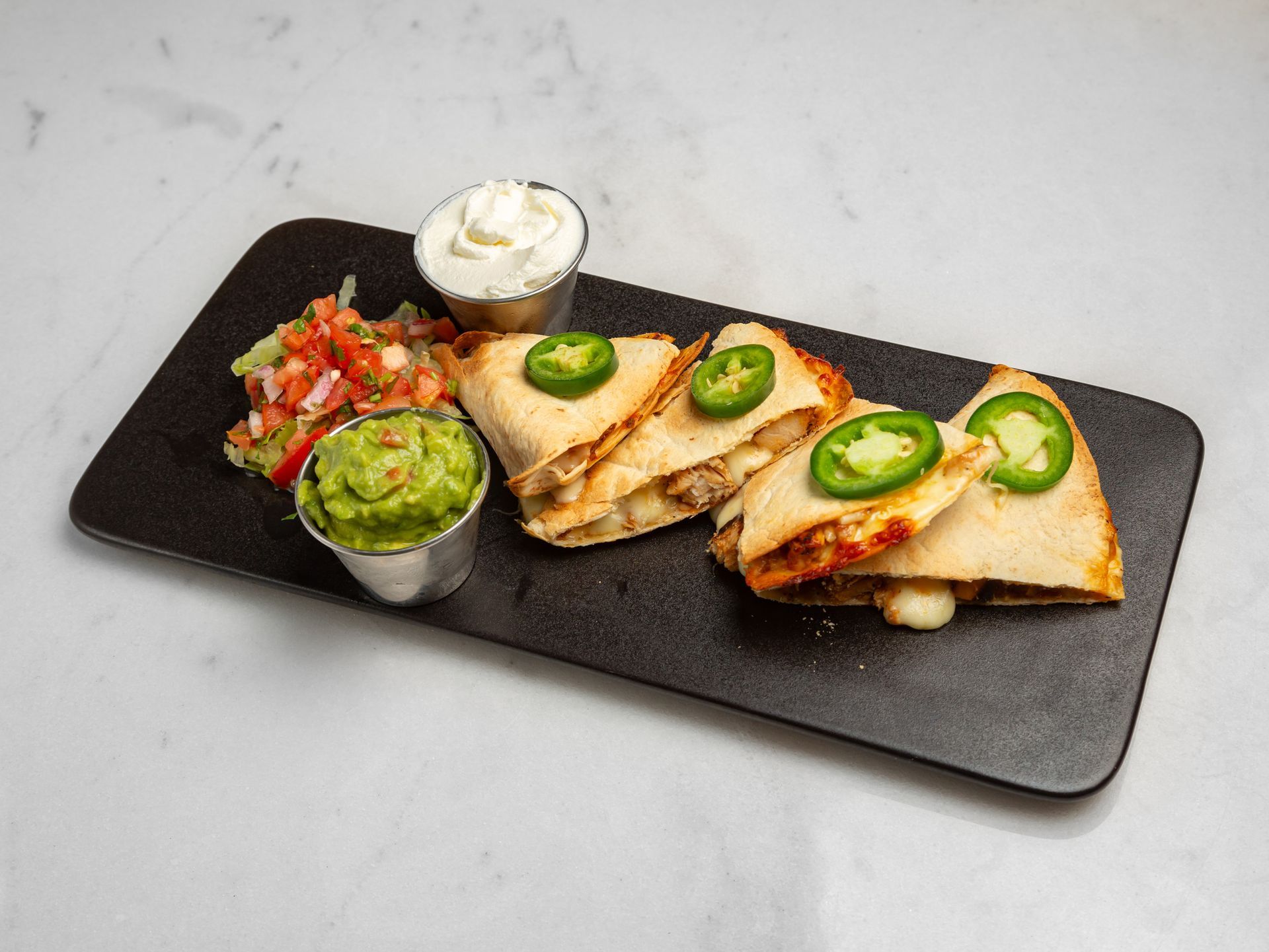 A black plate topped with quesadillas , guacamole and sour cream.