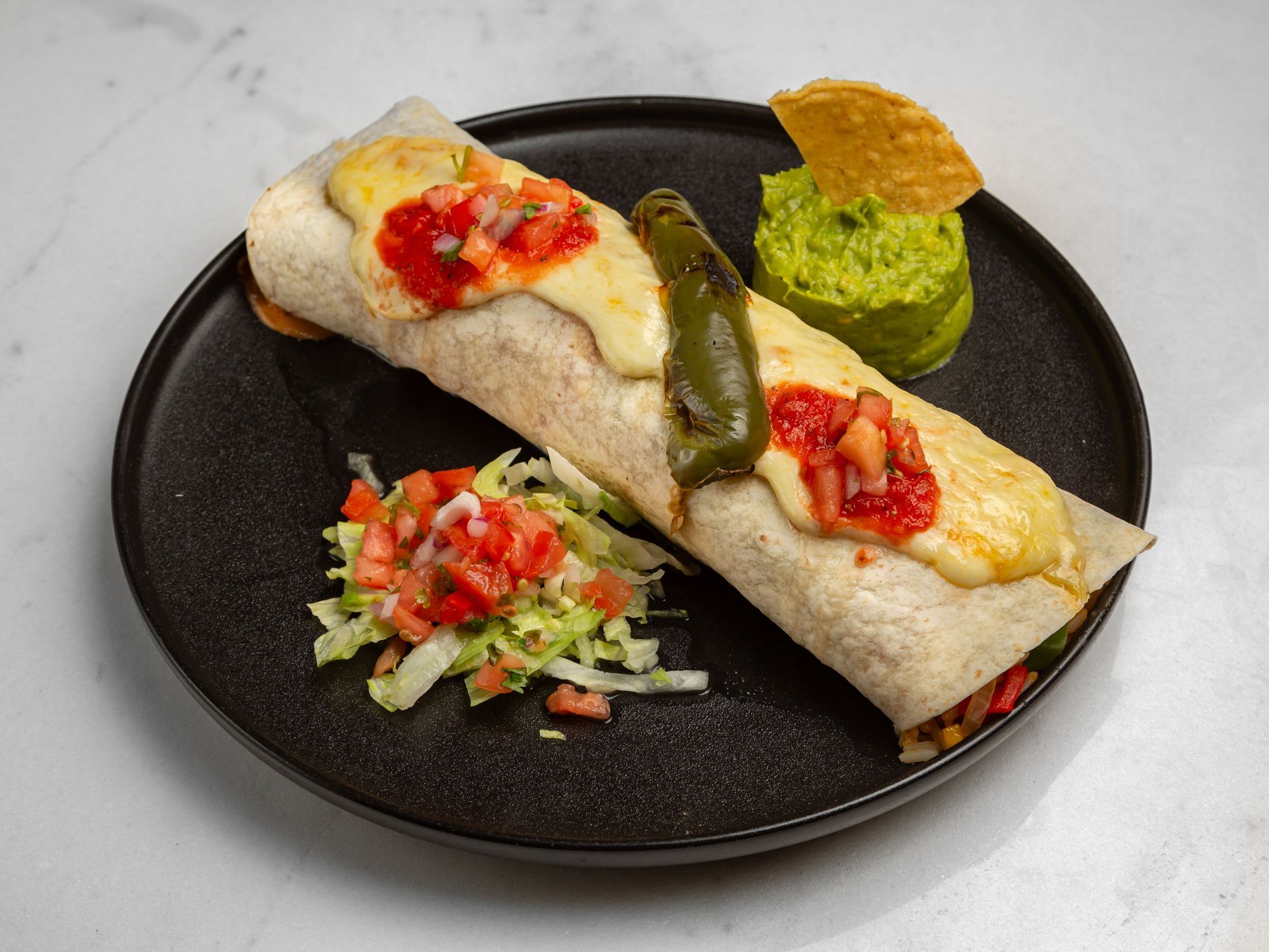 A burrito with cheese , tomatoes , lettuce and guacamole on a black plate.