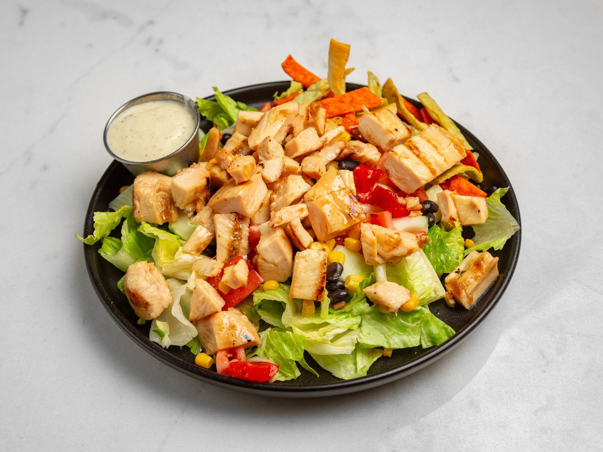 A salad with chicken , lettuce , carrots , black olives and ranch dressing on a black plate.