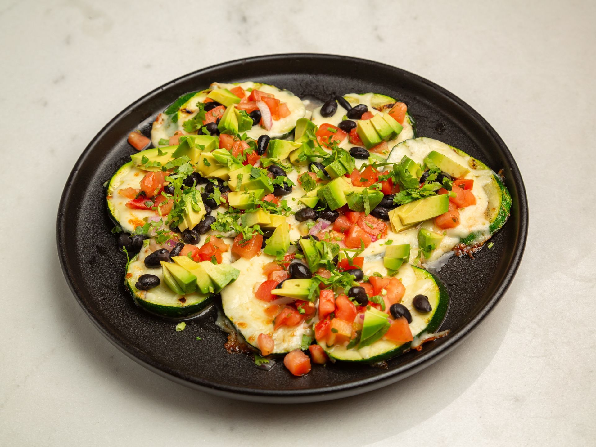 A black plate topped with zucchini , avocado , tomatoes and black beans.