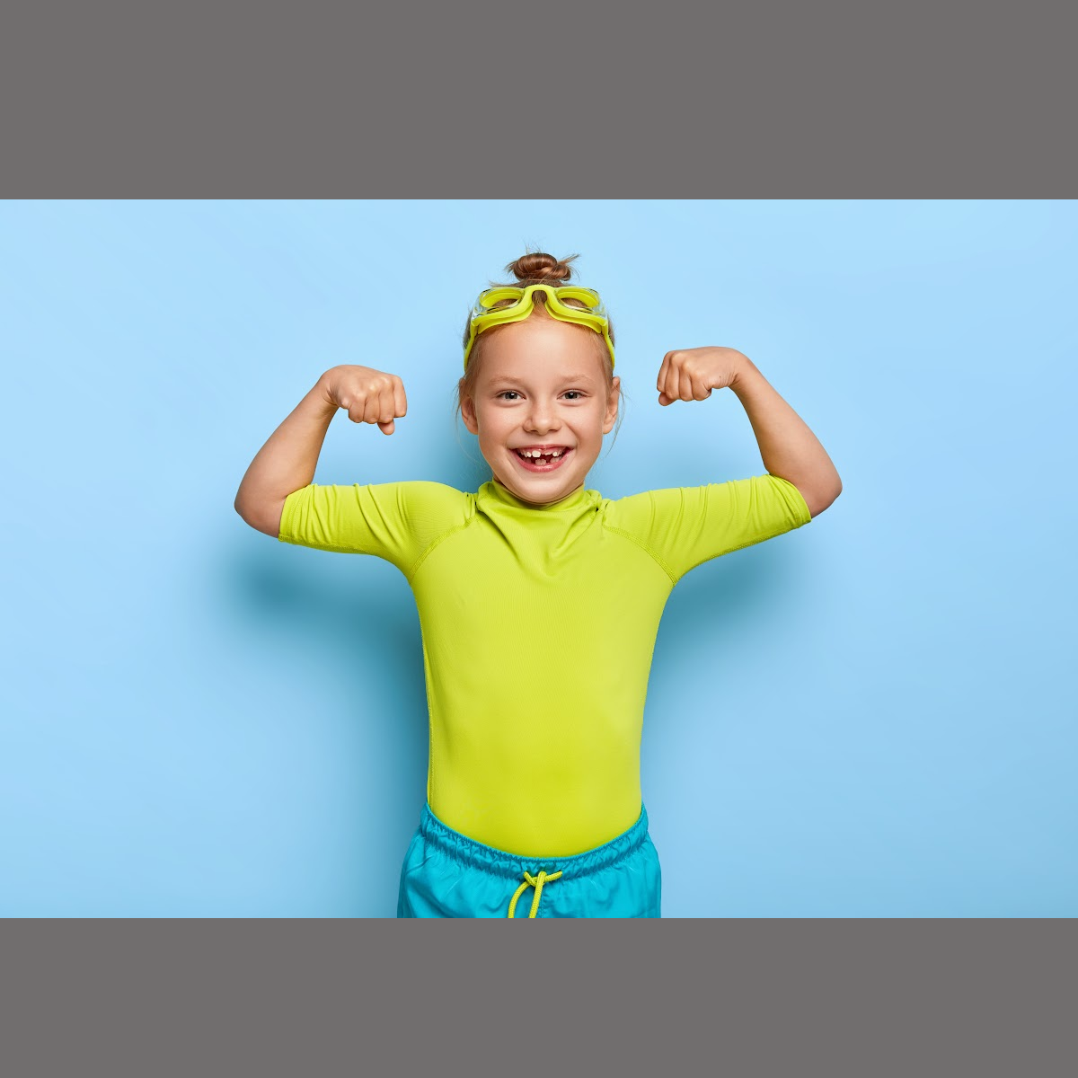 A young boy in a green shirt and blue shorts is flexing his muscles.