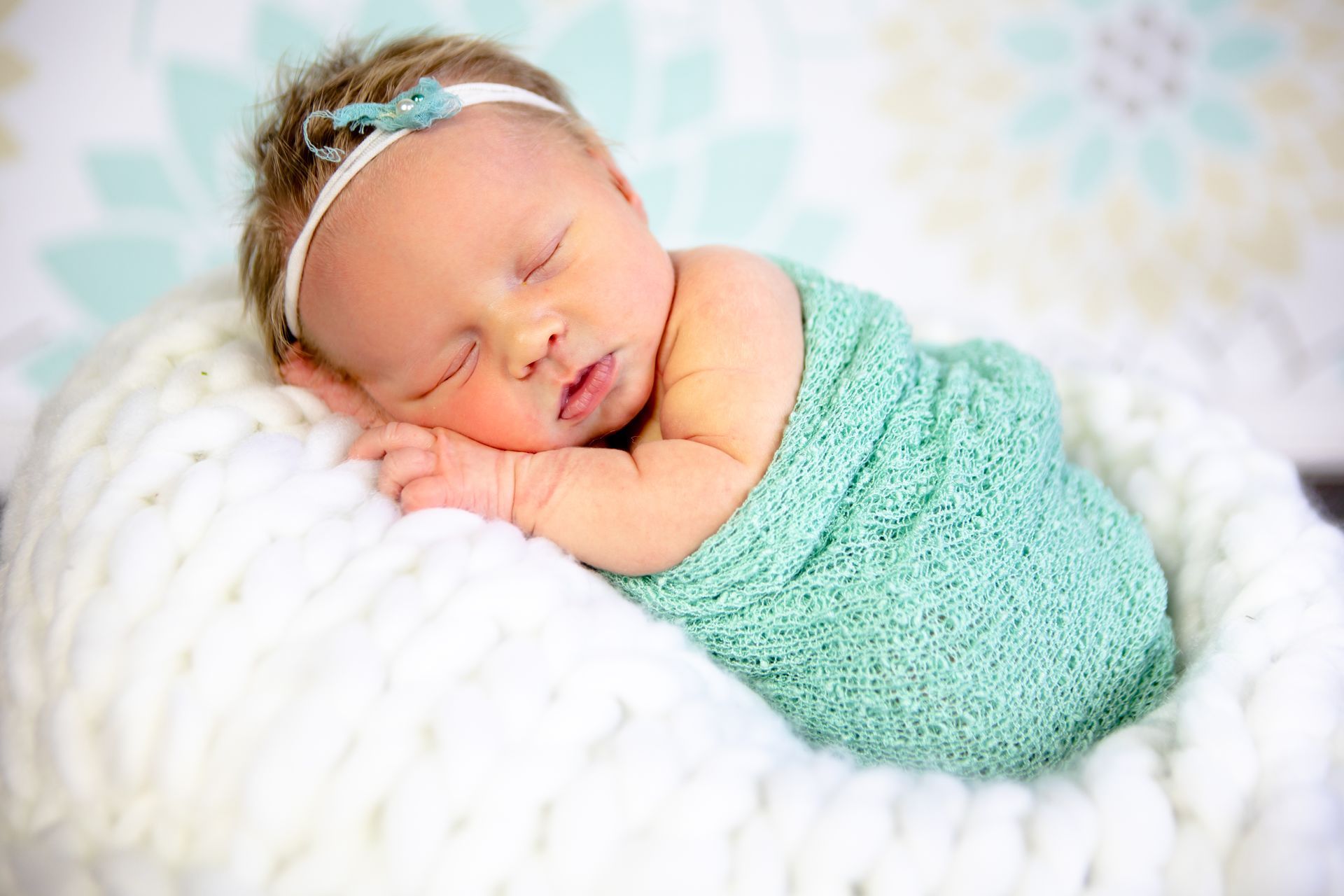 Newborn baby sleeping, wrapped in teal blanket, on white knitted blanket, wearing a headband.