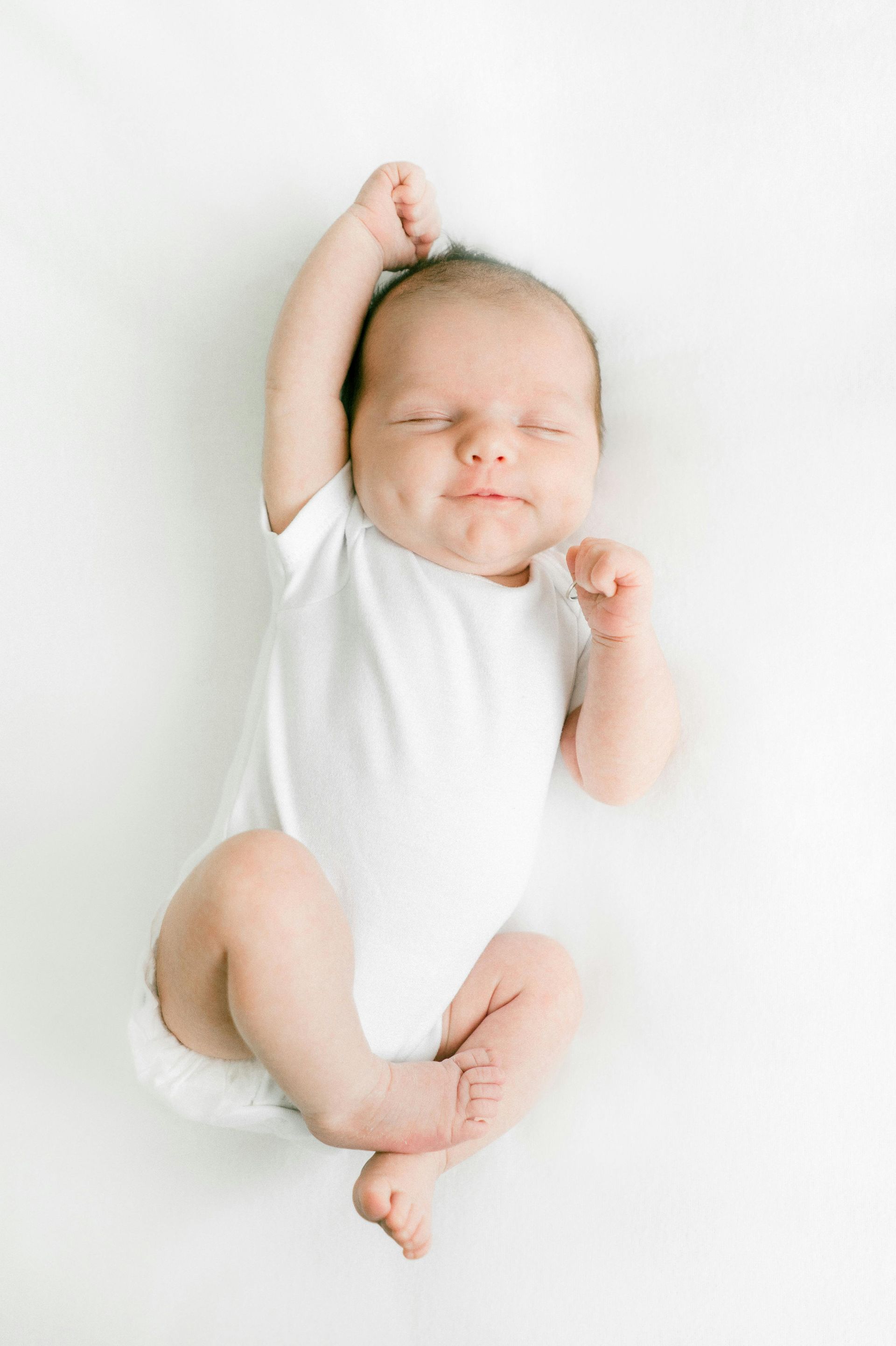 Newborn baby in white onesie, stretching, smiling, lying on white surface.