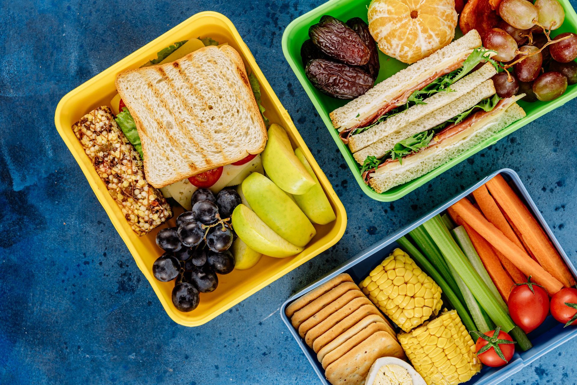 Three lunch boxes filled with sandwiches , fruit , vegetables and crackers.