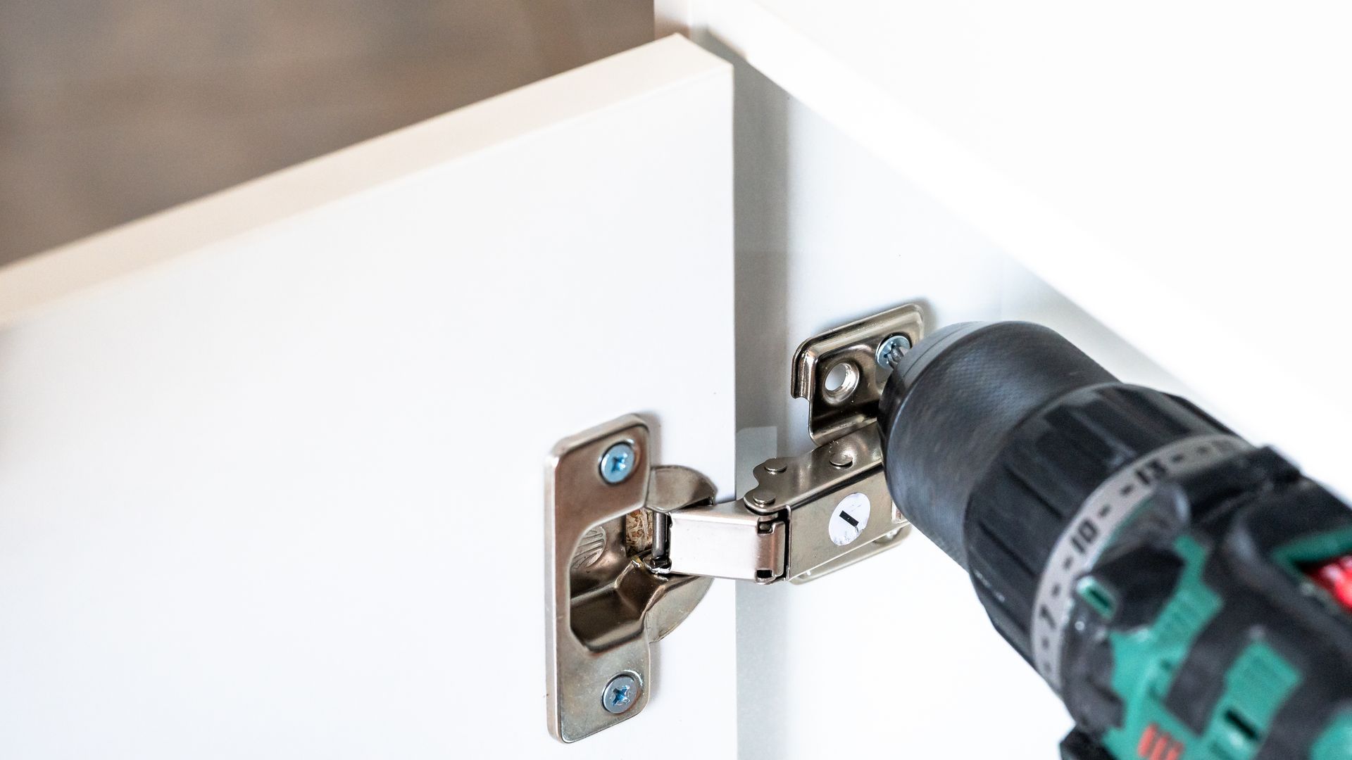 A cordless drill tightening a hinge on a white cabinet door