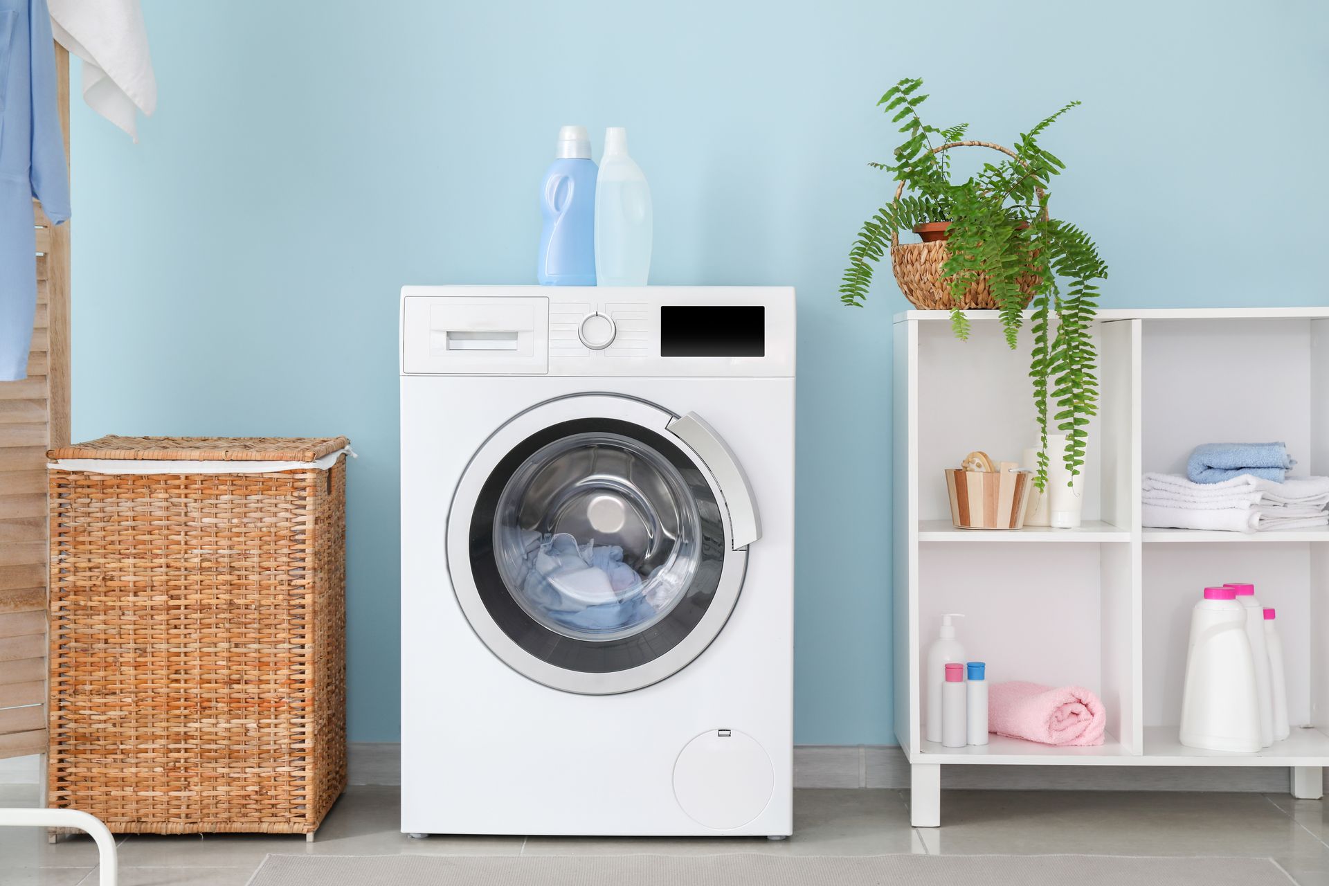 Laundry room with white front-loading washer, wicker hamper, shelf, and hanging fern.