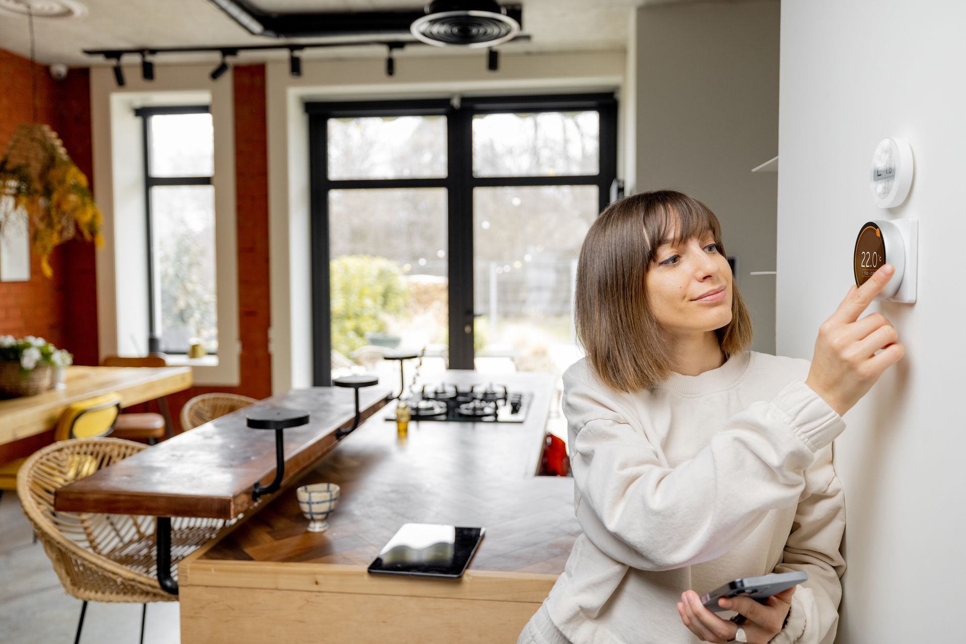 Woman adjusting a wall thermostat in a bright modern kitchen and living room.