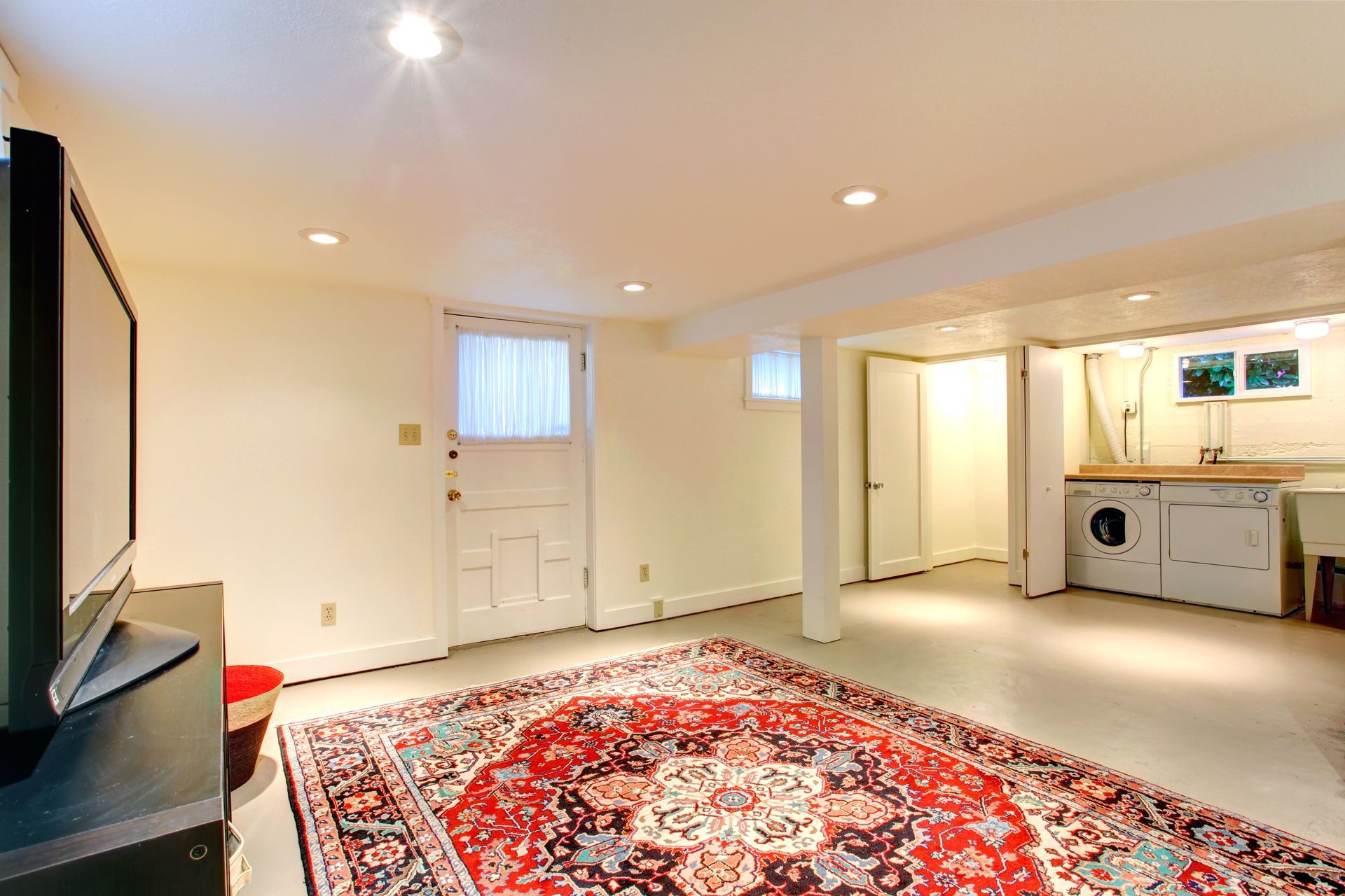 Empty basement rec room with red patterned rug, recessed lights, and a small kitchenette on the right
